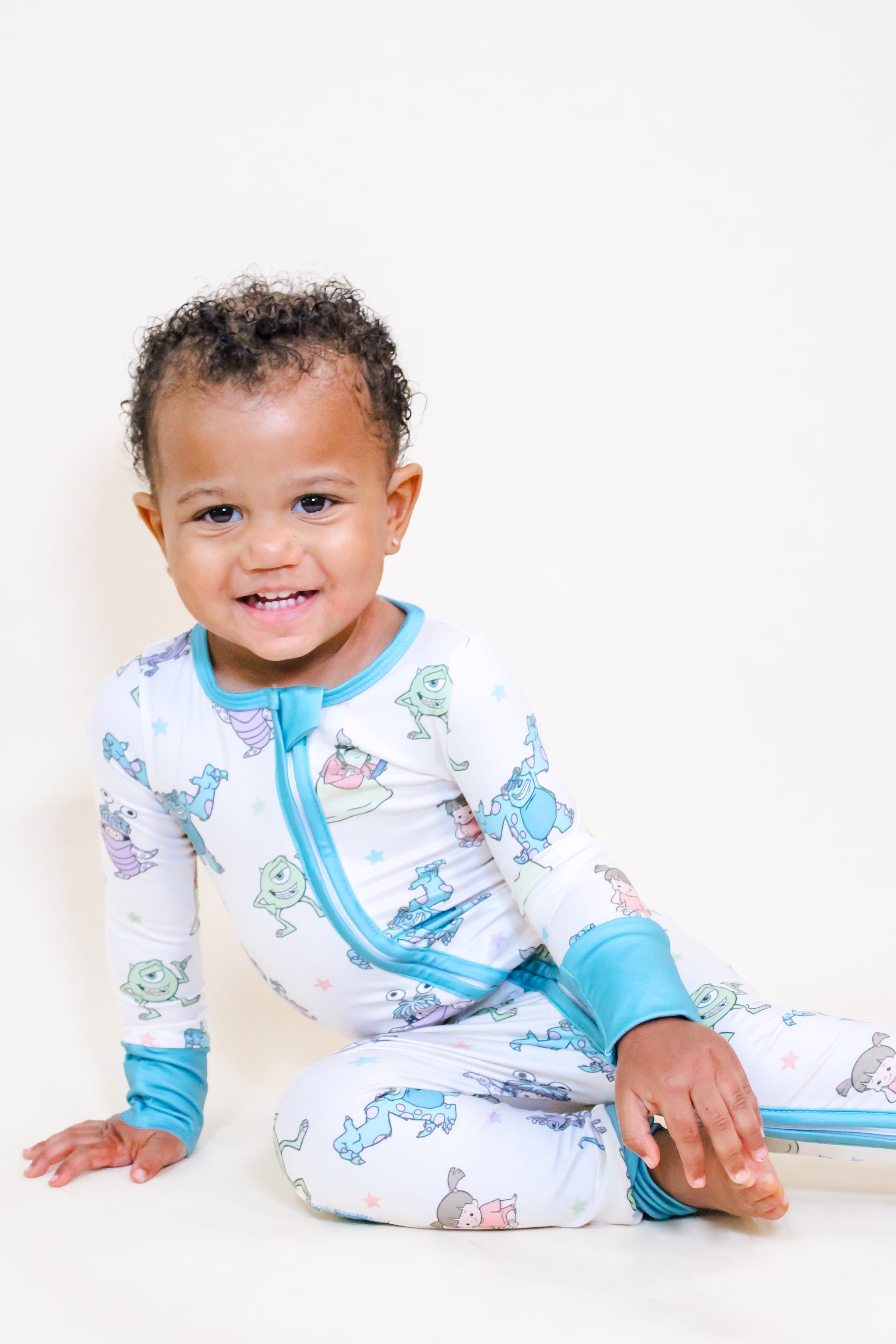 A smiling toddler with short curly hair wears the Tiny Tot Bundles Monster Pals Long Zippy, sitting on a light surface with one hand on the floor and legs stretched out.
