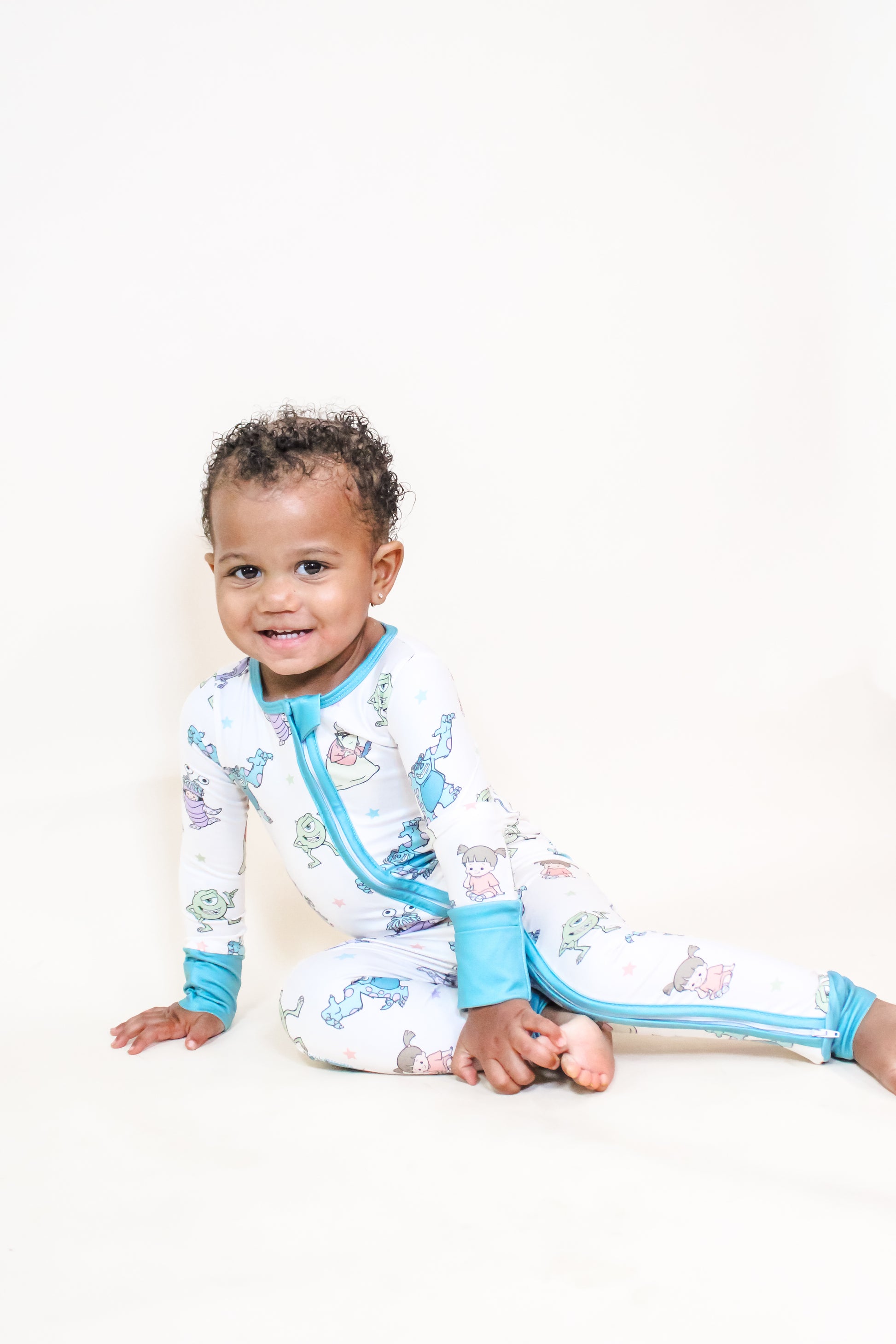 A smiling toddler with curly hair sits on the floor wearing the Tiny Tot Bundles Monster Pals Long Zippy, a white onesie featuring colorful cartoon animal prints and blue trim, against a plain light background.