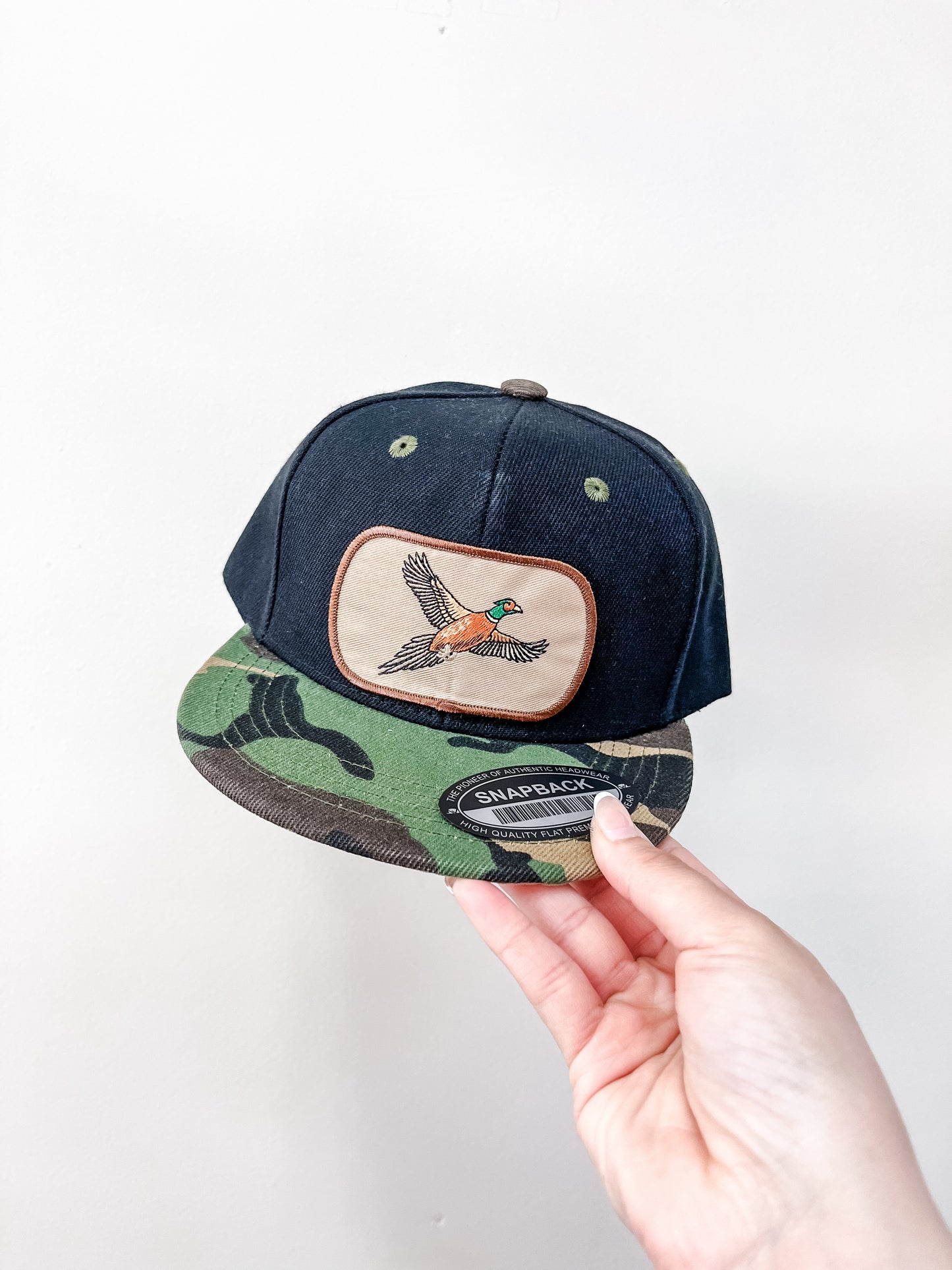 A hand holds the Thats Rad Upland Flight Hat, a black and camouflage snapback with an embroidered bird patch on the front panel, against a plain white wall.