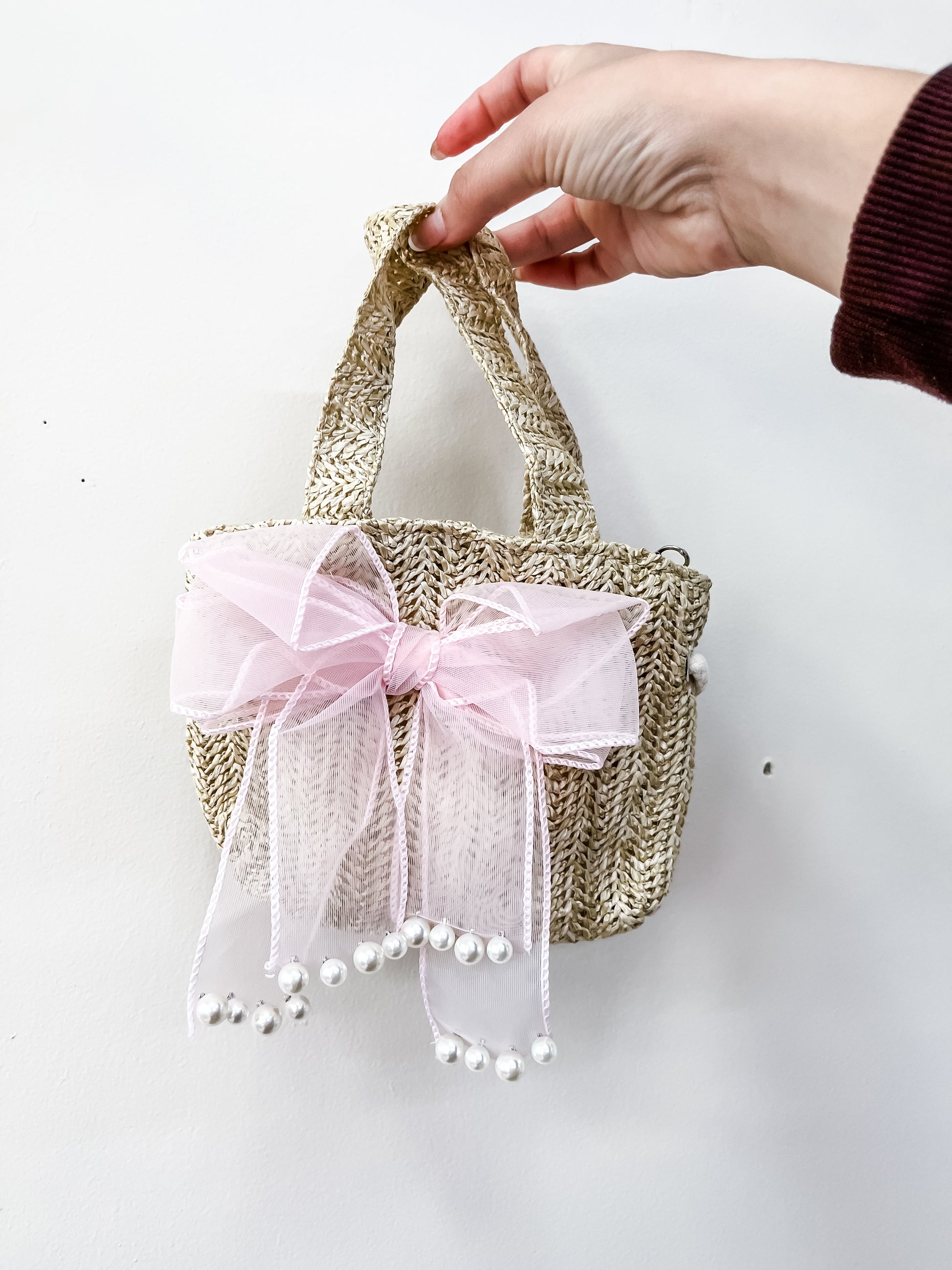 A hand holds the Tiny Tot Bundles Coquette Cutie Mini Bag, a small woven straw handbag adorned with a large pink tulle bow and dangling white pearls, set against a plain white background.