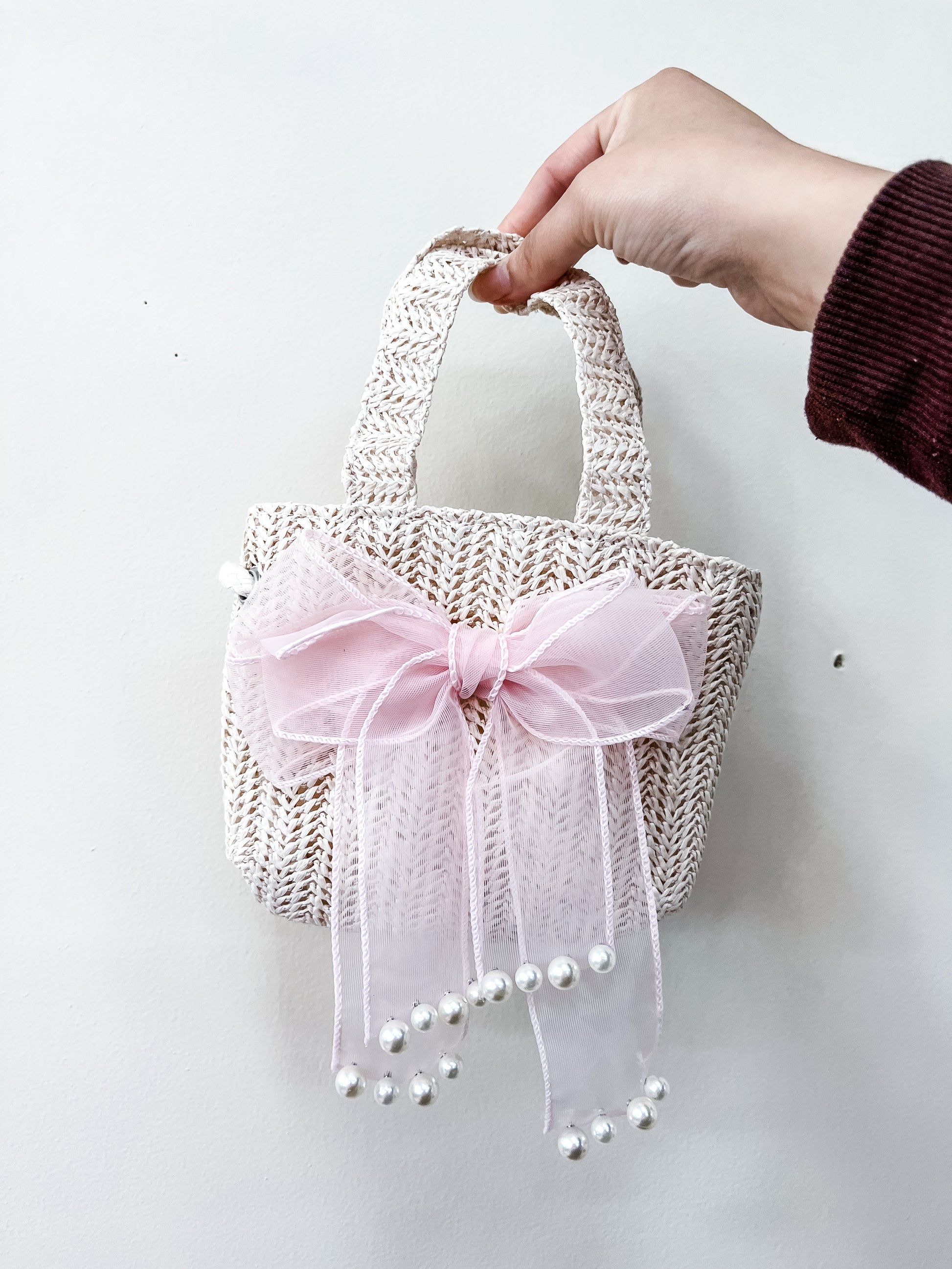 A hand holds the Tiny Tot Bundles Ivory Bow Mini Bag, a small woven straw handbag with a large pink tulle bow and dangling pearl-like beads, set against a plain white background.