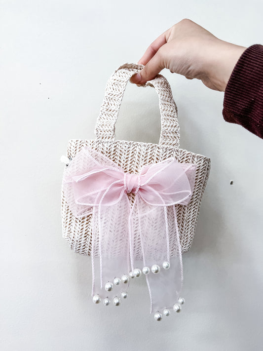 A hand holds the Tiny Tot Bundles Ivory Bow Mini Bag, a small woven straw handbag with a large pink tulle bow and dangling pearl-like beads, set against a plain white background.
