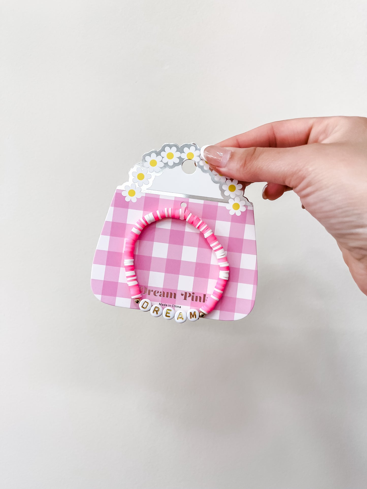 A hand holds the funteze Happy Words Bracelet, a pink and white beaded accessory with daisies and letter beads spelling "DREAM," displayed on a pink checkered card.