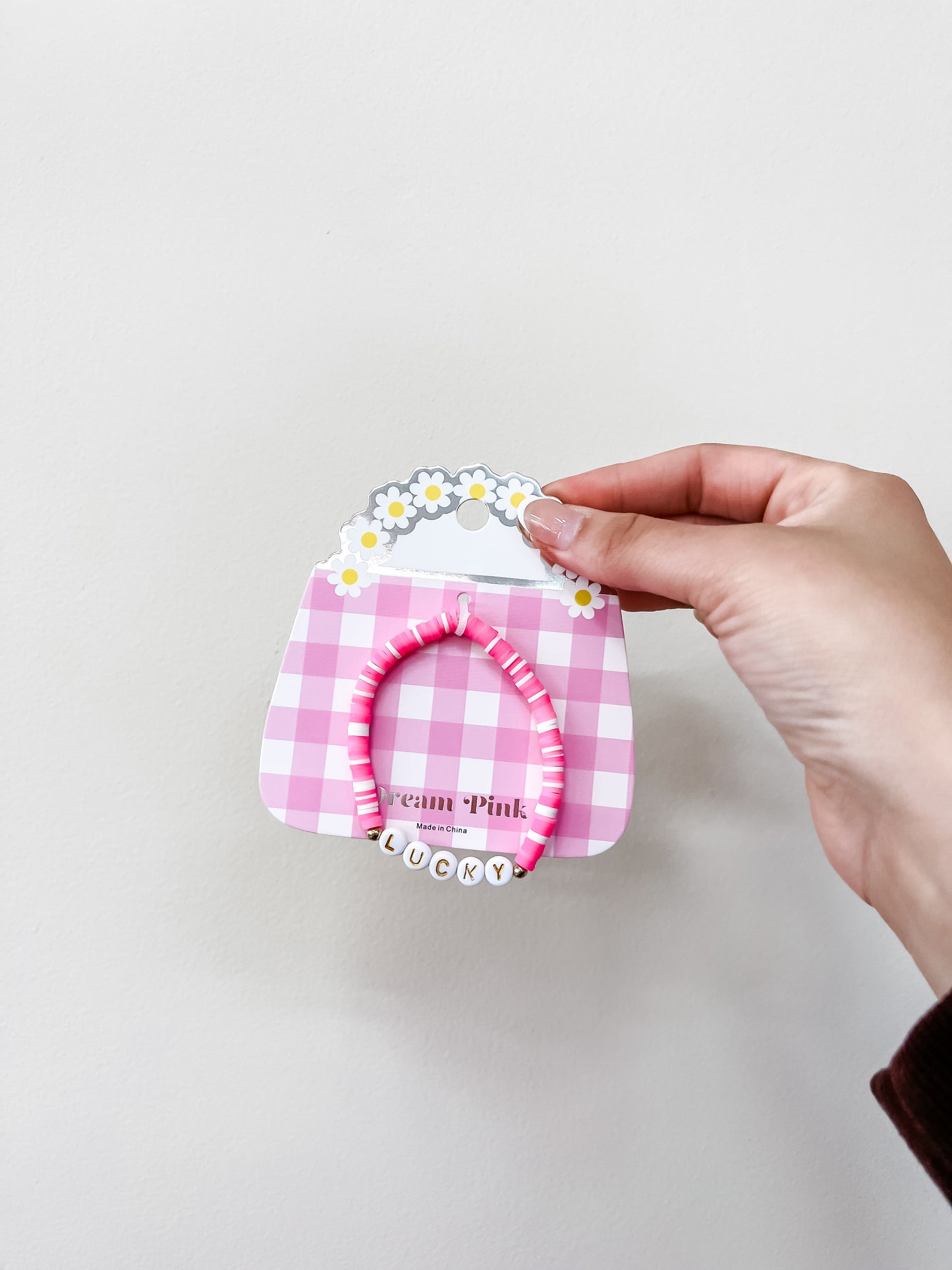 A hand holds a funteze Happy Words Bracelet with pink beads and alphabet letters spelling LUCKY, attached to a pink and white checkered purse-shaped card adorned with daisies.