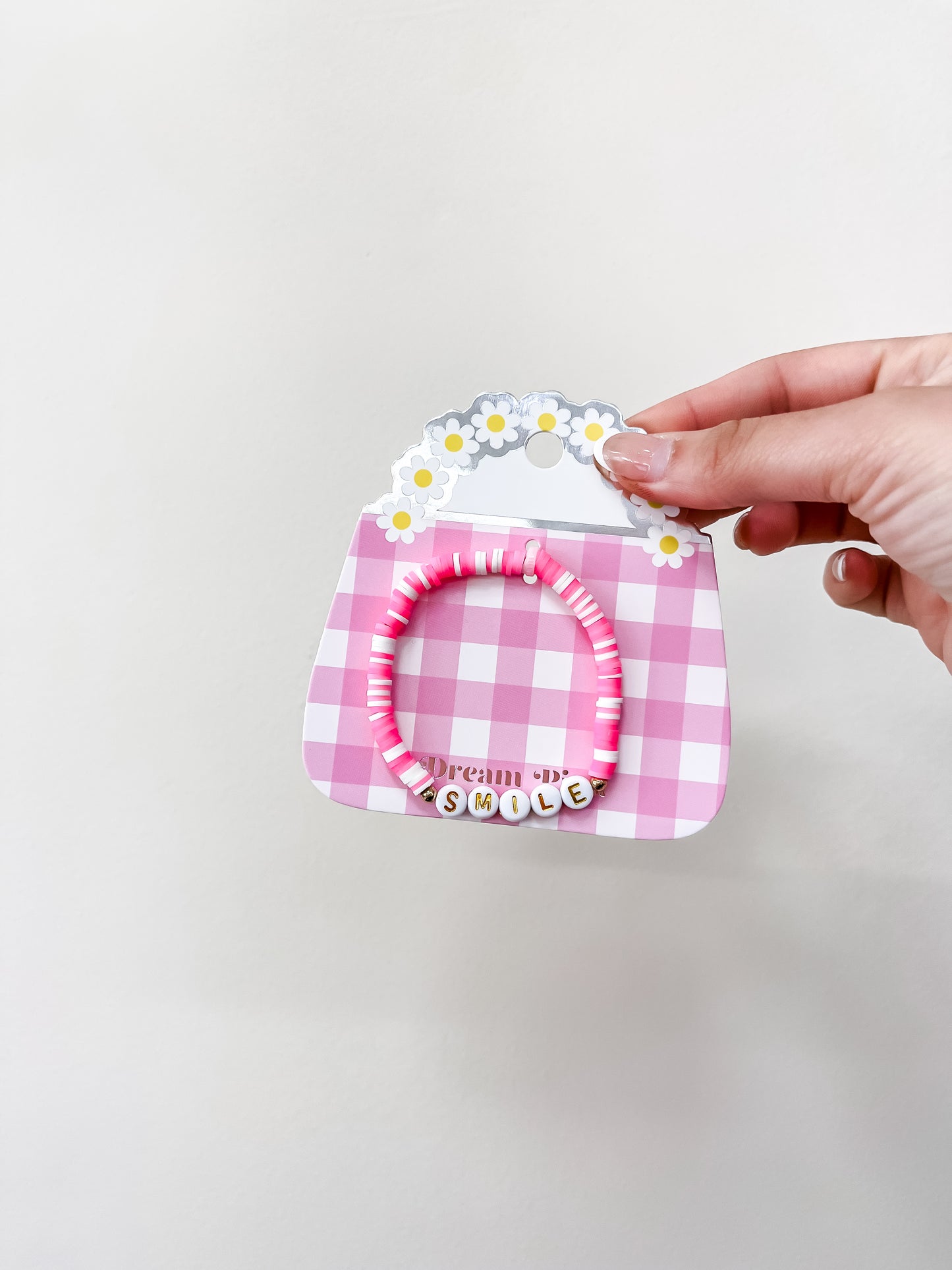 A hand holds a pink and white gingham card shaped like a purse with daisy details, showcasing funteze’s Happy Words Bracelet—a pink beaded bracelet that spells “SMILE” with letter beads.