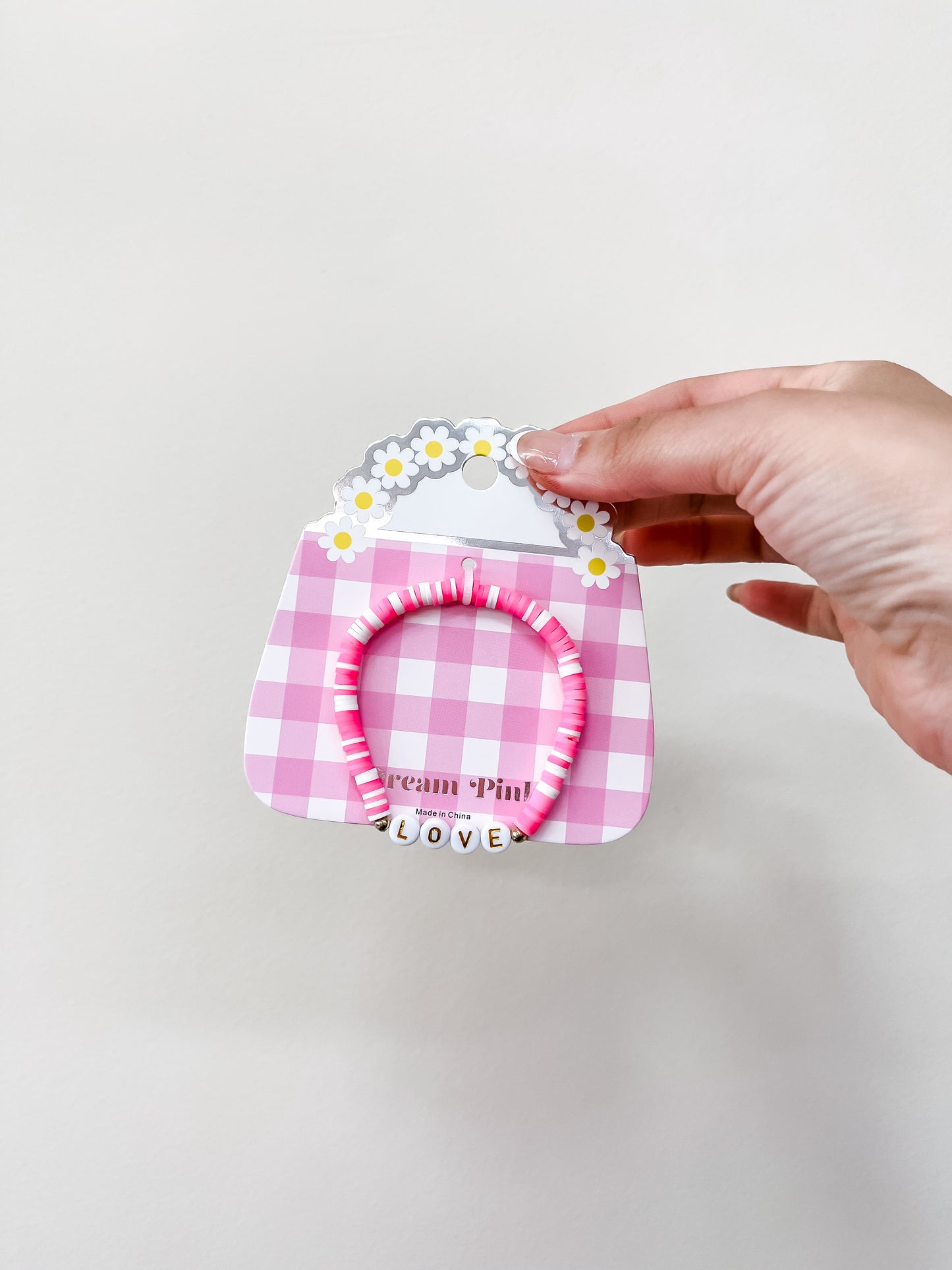 A hand holds the funteze Happy Words Bracelet on a pink and white checkered daisy card. The bracelet features pink beads and white letter beads spelling LOVE, shown against a plain, light background.