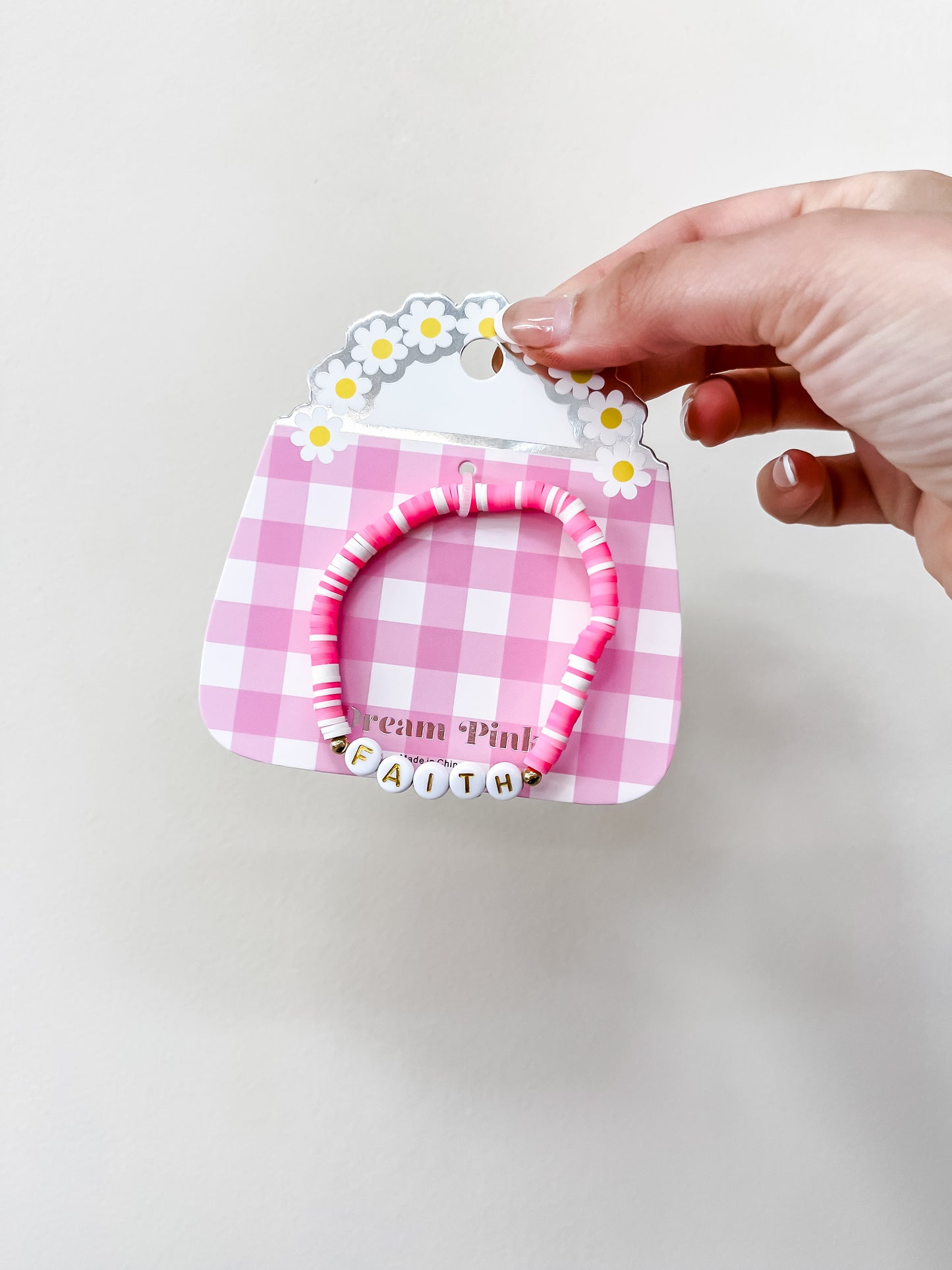 A hand holds the funteze Happy Words Bracelet, featuring pink and white beads and letter beads spelling FAITH, displayed on a pink gingham card with a floral handle.