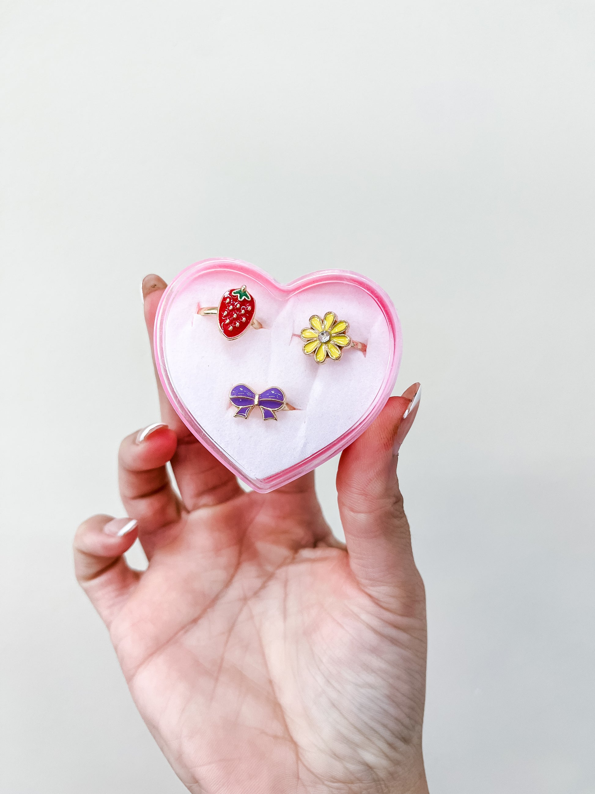 A manicured hand holds the Tiny Tot Bundles Sweetheart Ring Set: a pink, heart-shaped box with three rings—a strawberry, a yellow flower, and a purple bow.