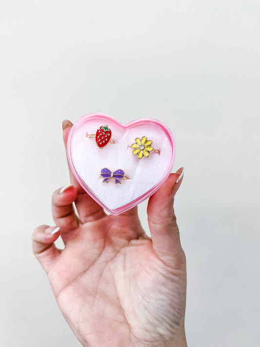 A manicured hand holds the Tiny Tot Bundles Sweetheart Ring Set: a pink, heart-shaped box with three rings—a strawberry, a yellow flower, and a purple bow.