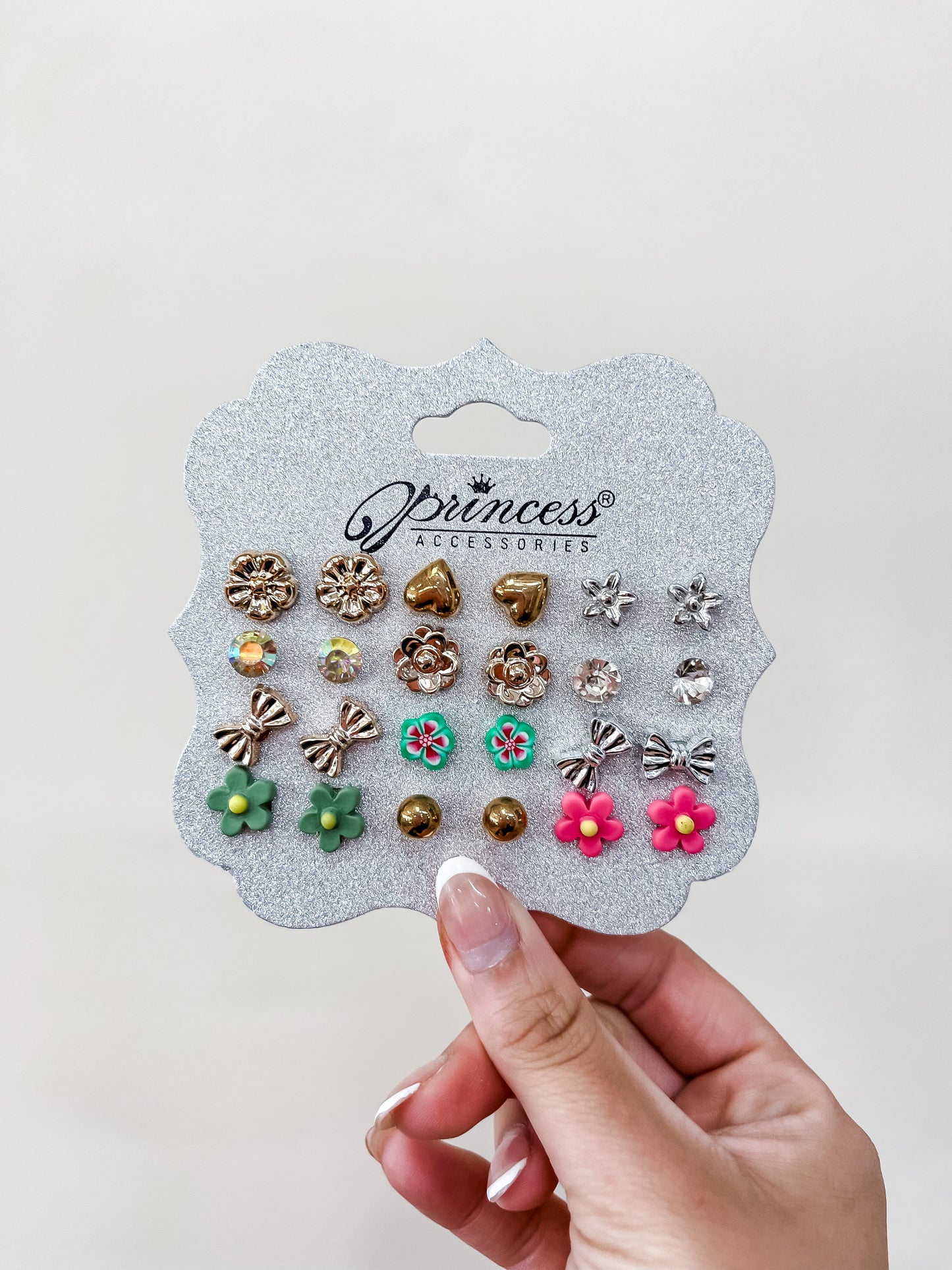 A hand holds a card labeled "Princess Earing Set" by funteze, showing 18 pairs of earrings with floral, heart, bow, and round designs in gold, pink, green, and clear colors against a plain background.