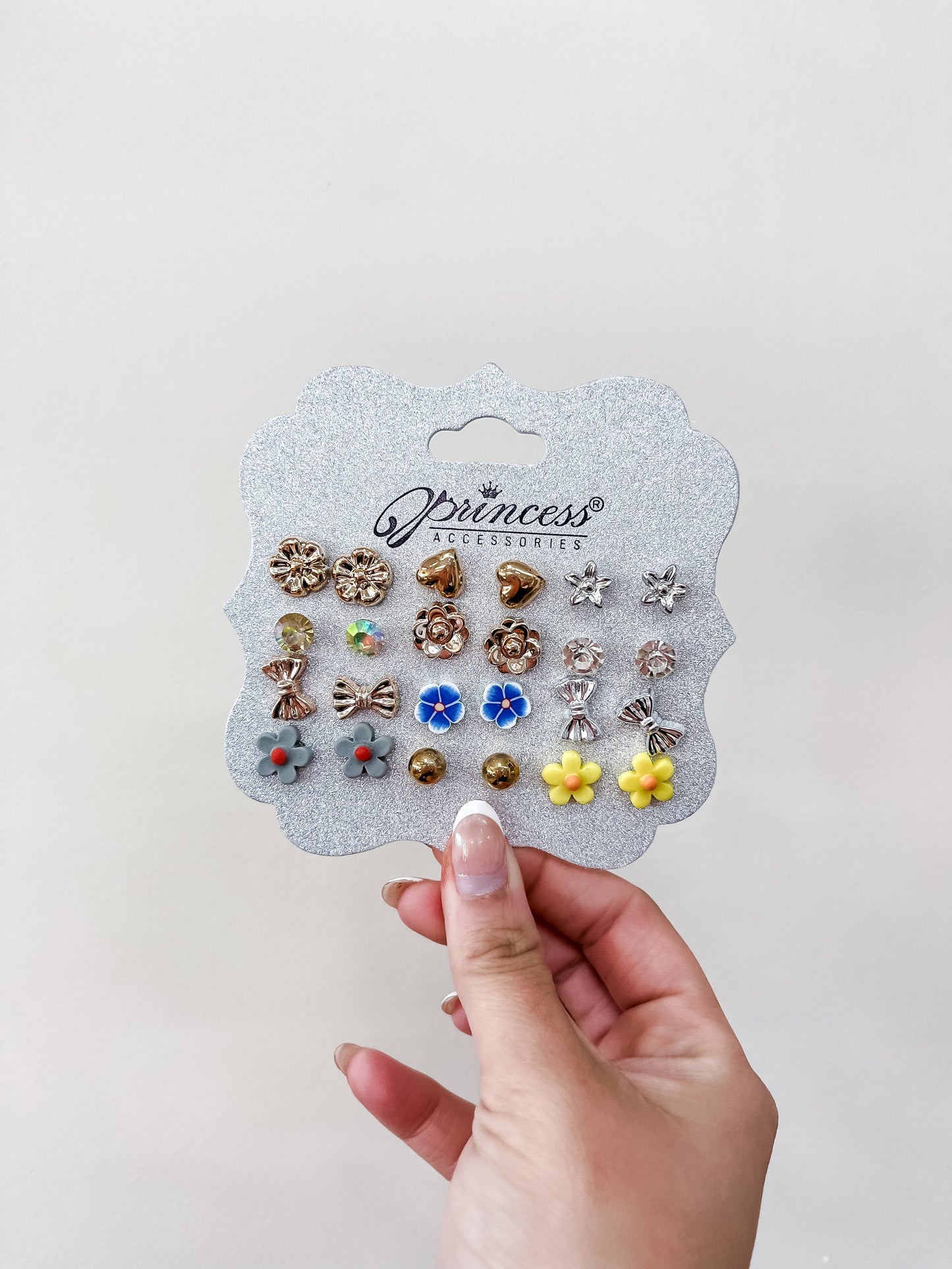 A hand holds a card labeled "funteze Princess Earing Set," showing 12 pairs of colorful stud earrings with flower and metallic designs, against a plain light background.