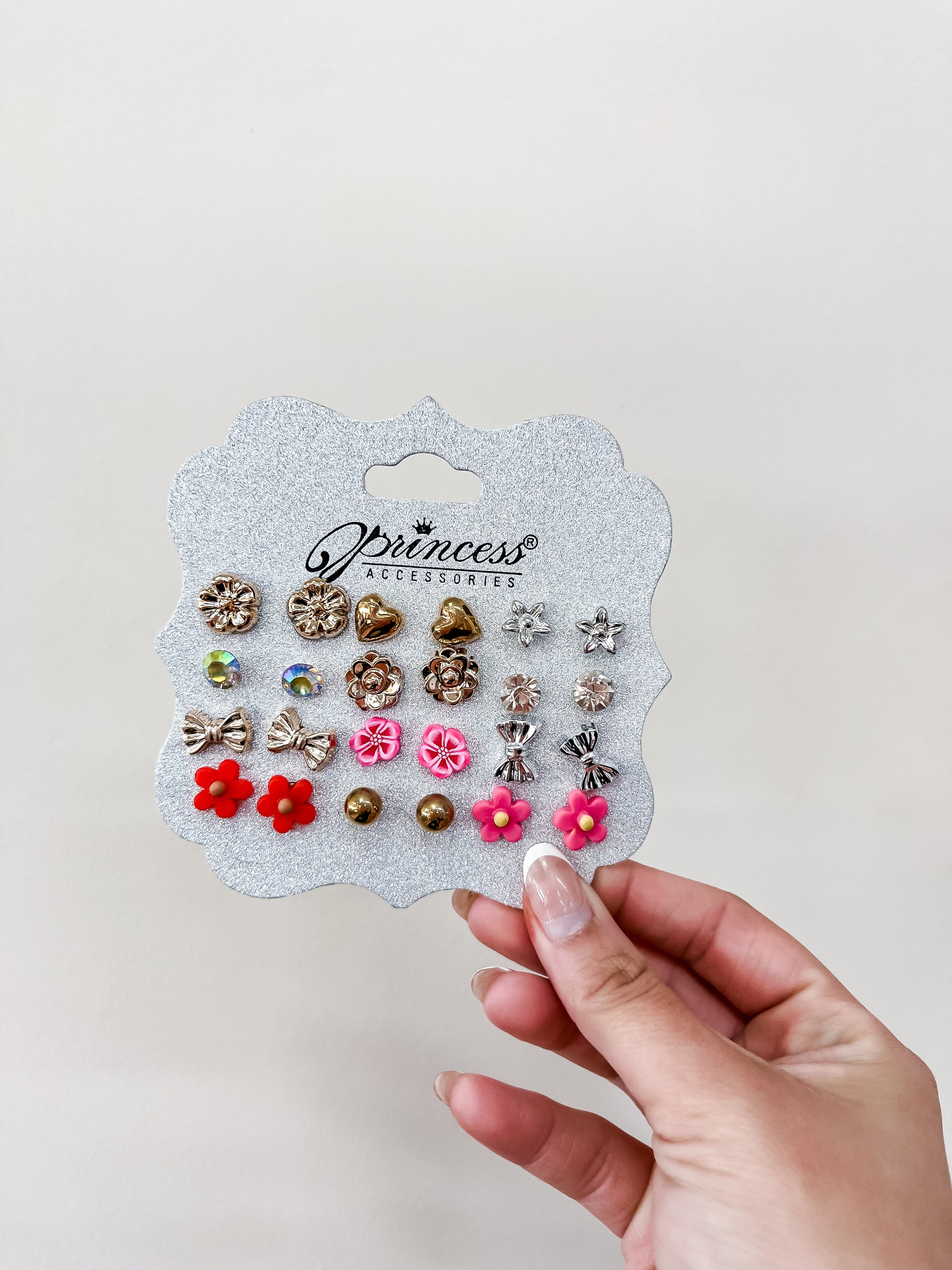 A hand holds a silver glittery card labeled "Princess Earing Set" by funteze, featuring 12 pairs of colorful stud earrings with designs like flowers, bows, and gems against a plain light background.
