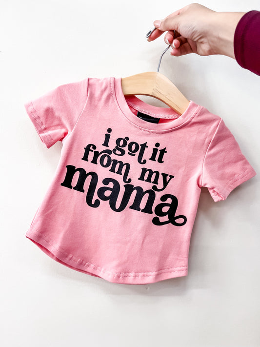 A hand holds a wooden hanger with the Beauts & Beaus Got It From Mama Mauve T-Shirt, featuring the phrase “i got it from my mama” in bold black letters on the front.