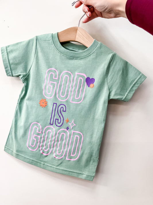 A hand holds up a green Beauts & Beaus God Is Good T-Shirt for toddlers on a wooden hanger. The shirt features "GOD IS GOOD" in bold, colorful letters with flowers, hearts, and sparkles.