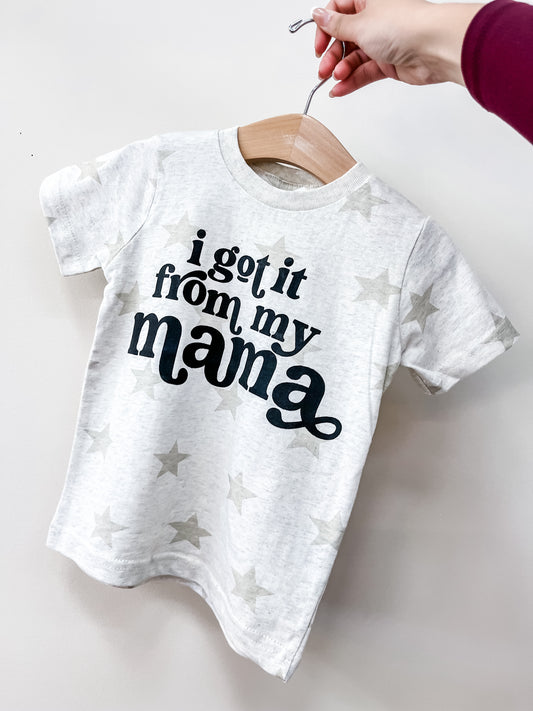 A hand holds up the Beauts & Beaus Got It from Mama Star T-Shirt, a light gray children’s short-sleeve tee with a star pattern and bold black text that says, "I got it from my mama.