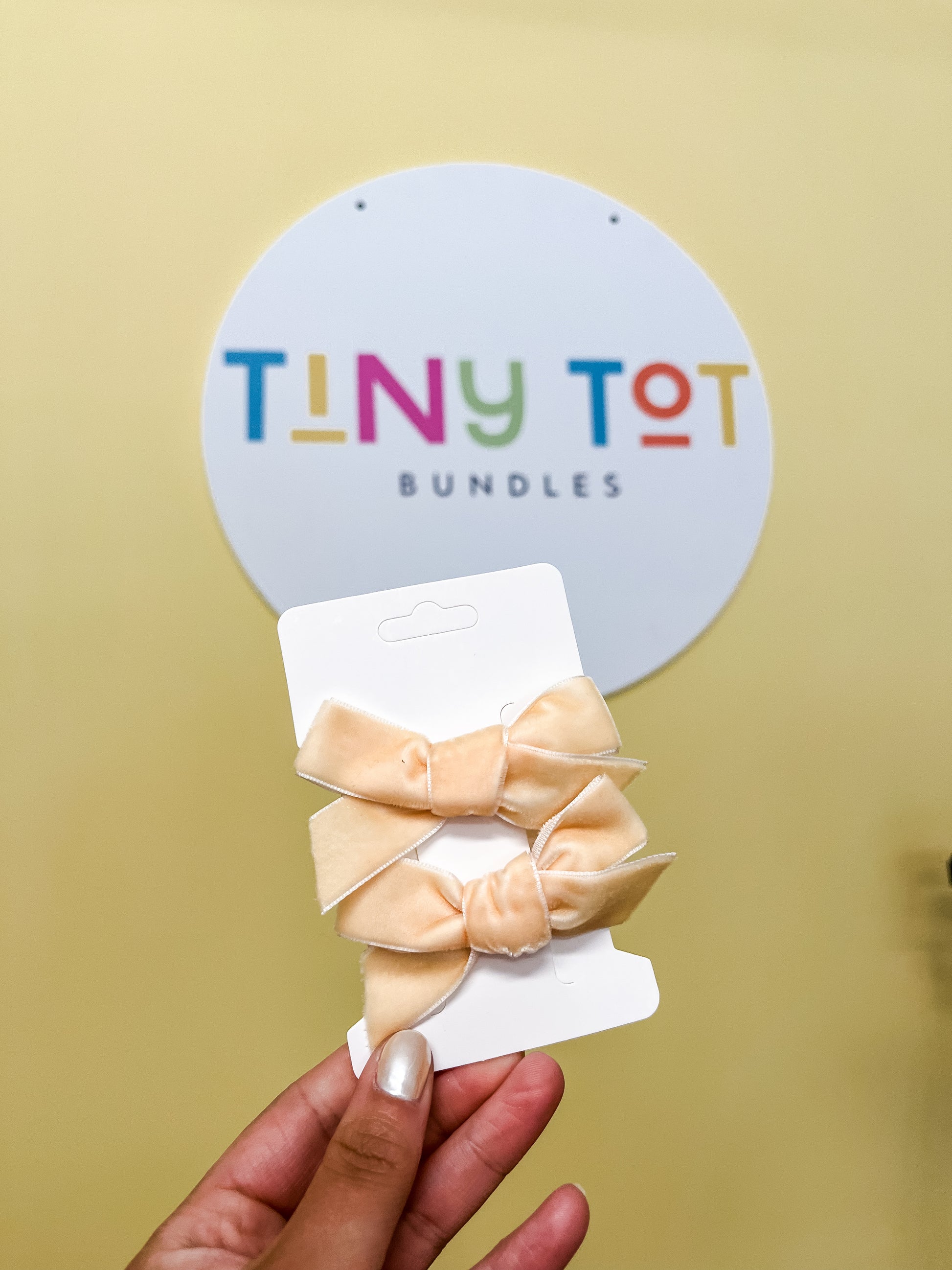 A hand holds a white card displaying two light peach Velvet Luxe Bows from Tiny Tot Bundles in front of a sign that reads "TINY TOT BUNDLES" in colorful letters on a white circle.