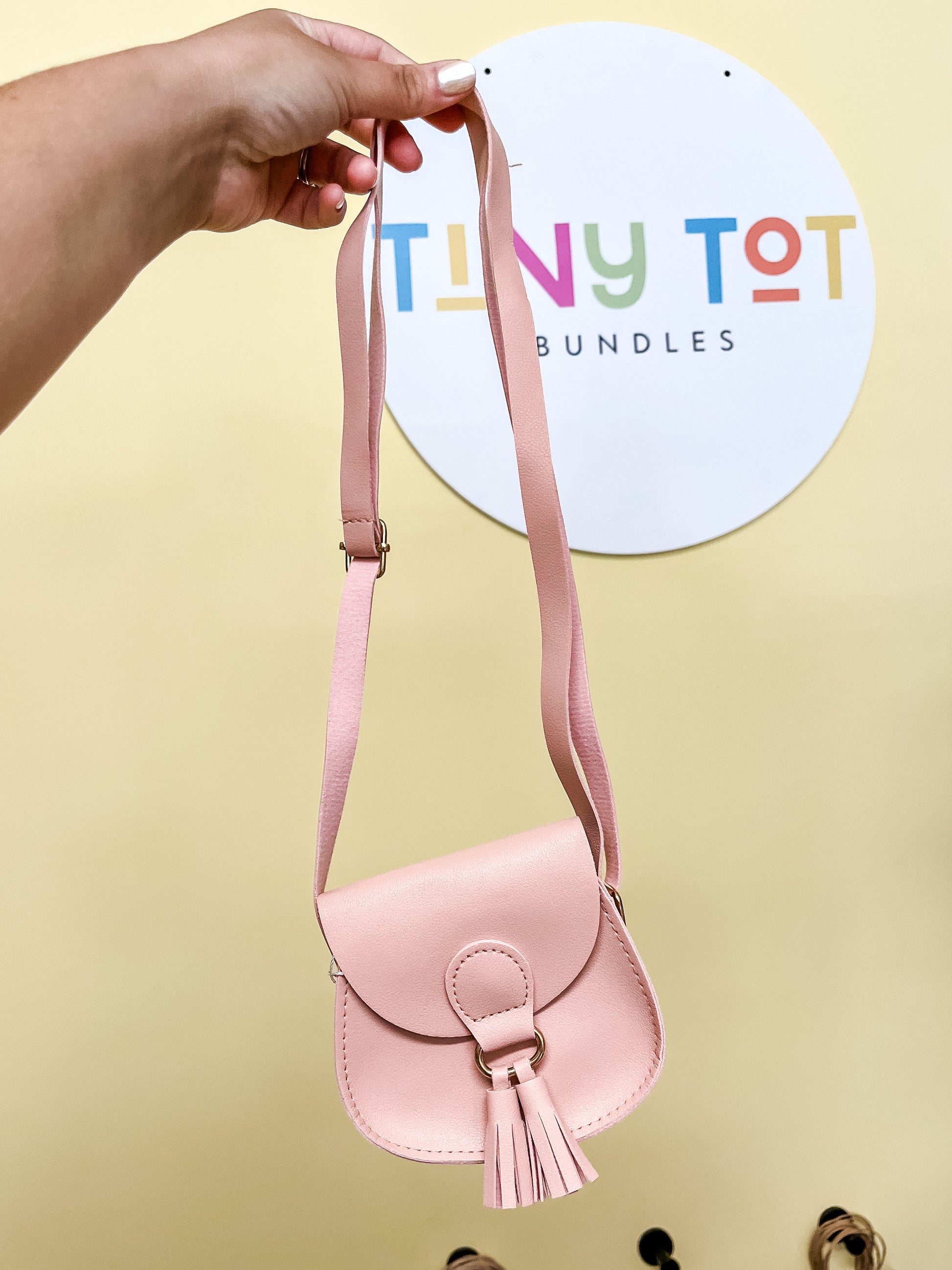 A hand holds the Daisy Coin Purse, a small, light pink accessory by Tiny Tot Bundles, in front of a circular sign with the brand's colorful logo.