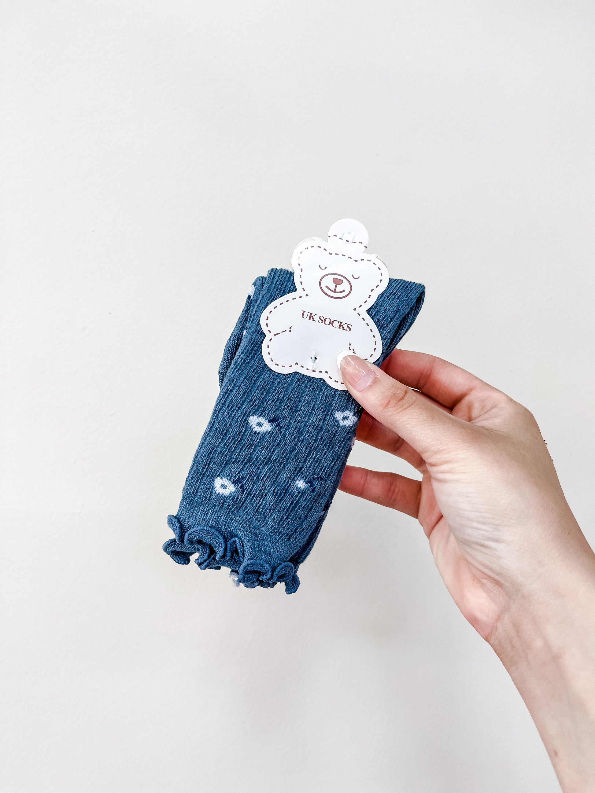 A hand holds a pair of Blue Petal Socks by Tiny Tot Bundles, featuring blue ruffles and small white patterns, packaged with a bear-shaped tag, against a plain light background.