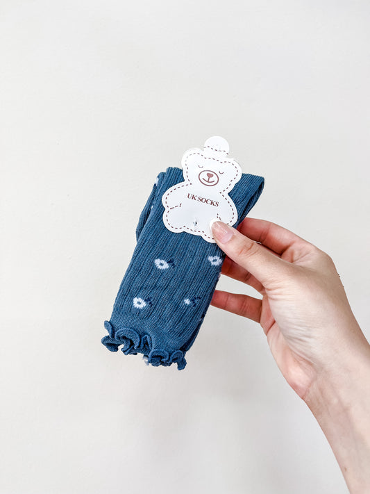 A hand holds a pair of Blue Petal Socks by Tiny Tot Bundles, featuring blue ruffles and small white patterns, packaged with a bear-shaped tag, against a plain light background.