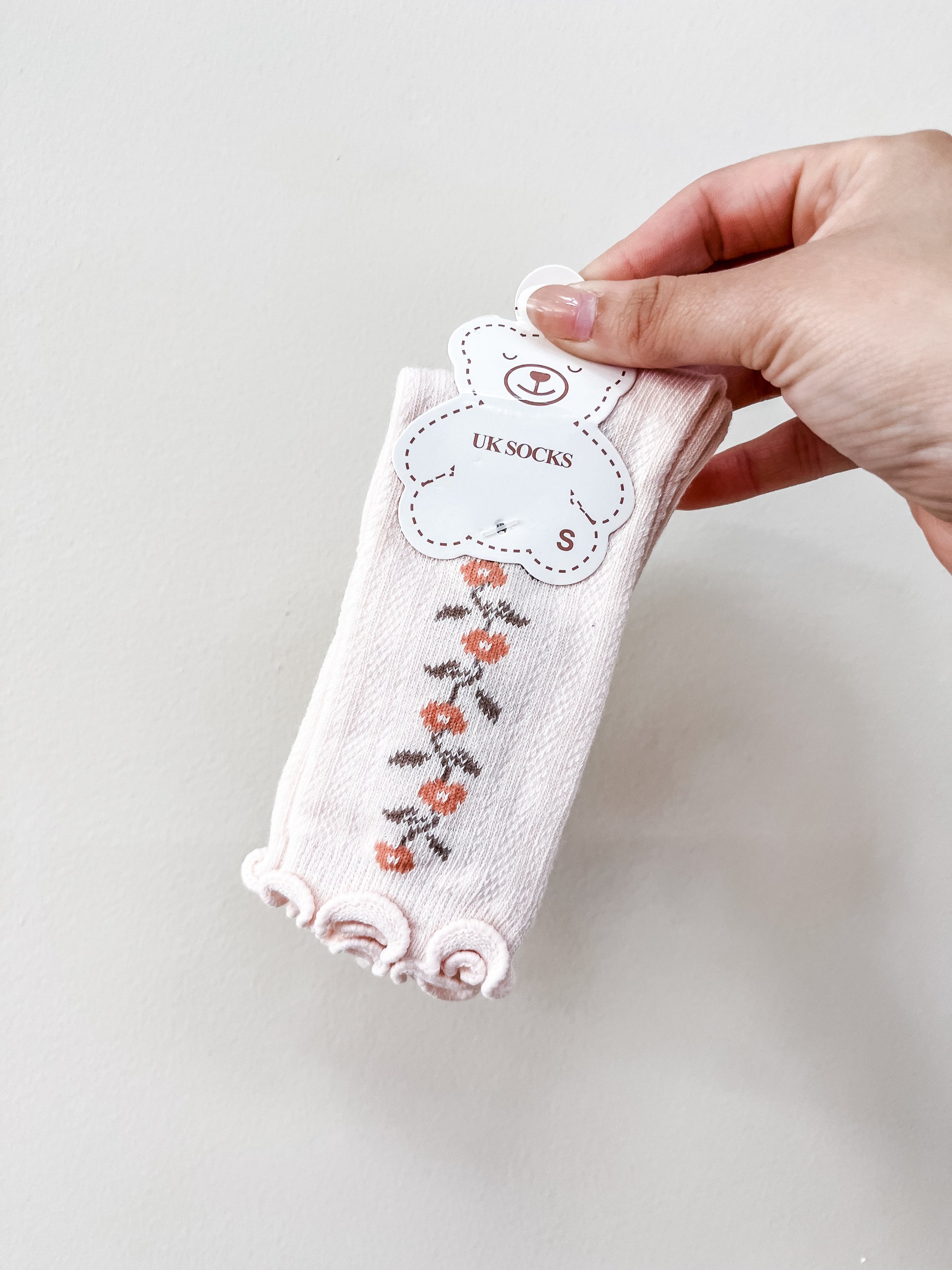 A hand holds a pair of Tiny Tot Bundles Pink Petal Socks featuring a floral pattern and frilled edges, labeled with a bear-shaped tag.