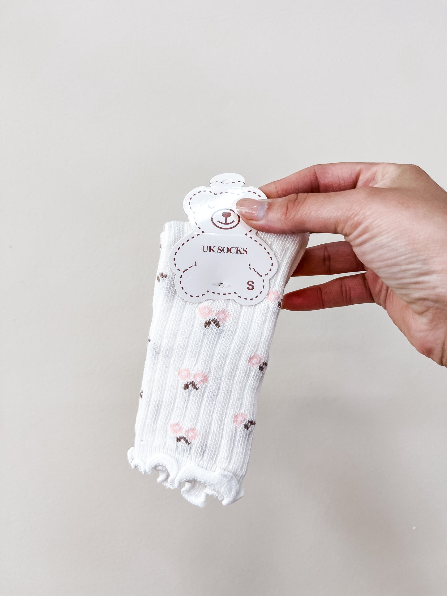 A hand holds a pair of Ivory Petal Socks by Tiny Tot Bundles, featuring white fabric with pink floral patterns and frilled edges, against a plain light background.