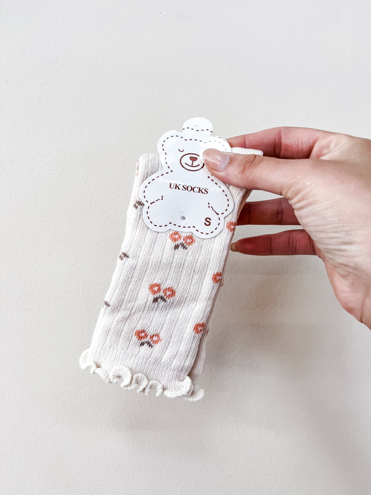 A hand holds Tiny Tot Bundles’ Beige Petal Socks, featuring small orange flowers and frilled edges, packaged with a bear-shaped tag labeled “UK SOCKS S.”.