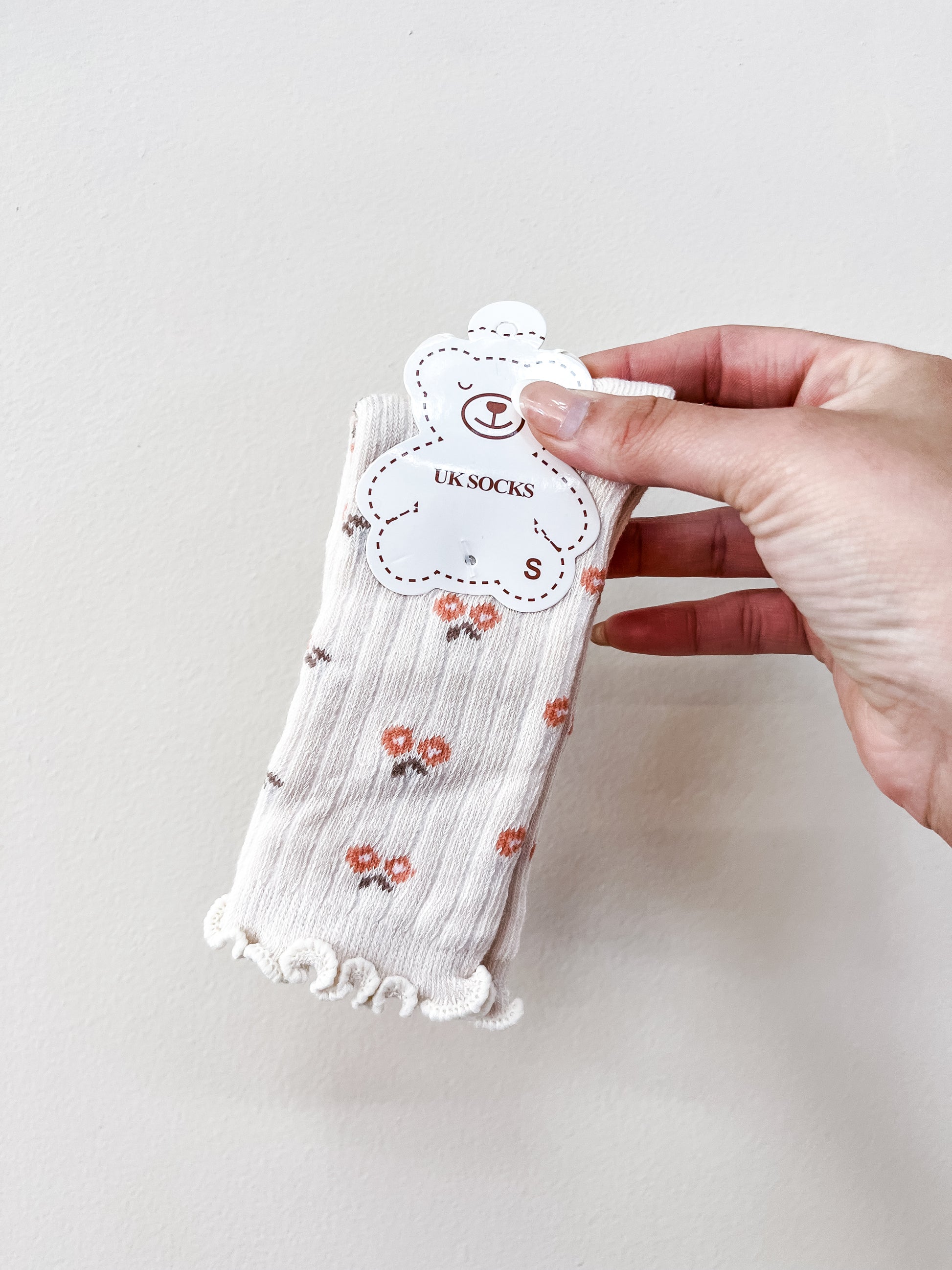 A hand holds Tiny Tot Bundles’ Beige Petal Socks, featuring small orange flowers and frilled edges, packaged with a bear-shaped tag labeled “UK SOCKS S.”.