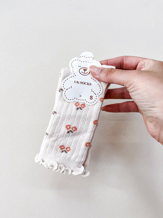 A hand holds Tiny Tot Bundles’ Beige Petal Socks, featuring small orange flowers and frilled edges, packaged with a bear-shaped tag labeled “UK SOCKS S.”.