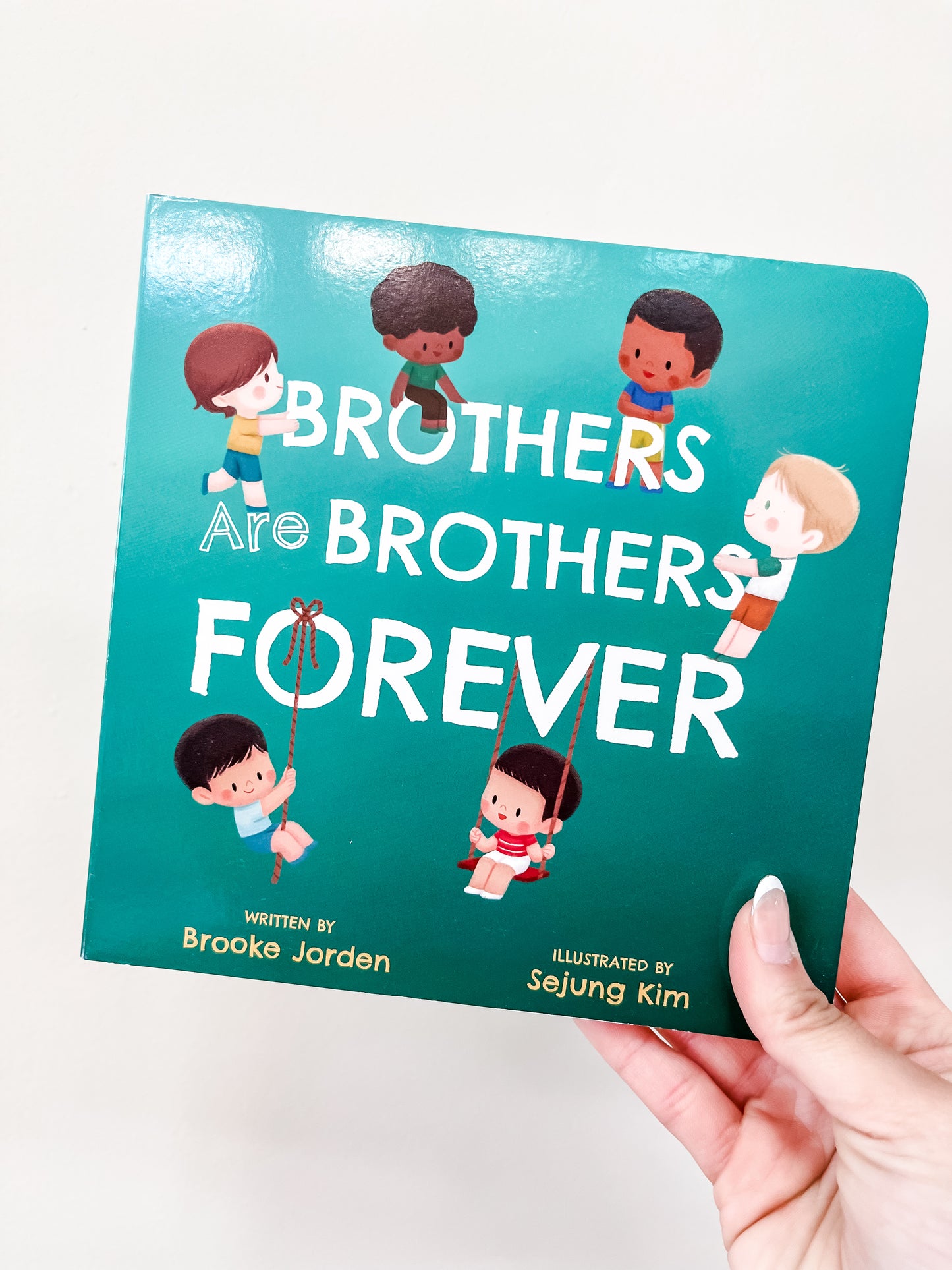 A hand holds "Brothers Are Brothers Forever" by familuis, featuring illustrated boys playing on a teal cover.