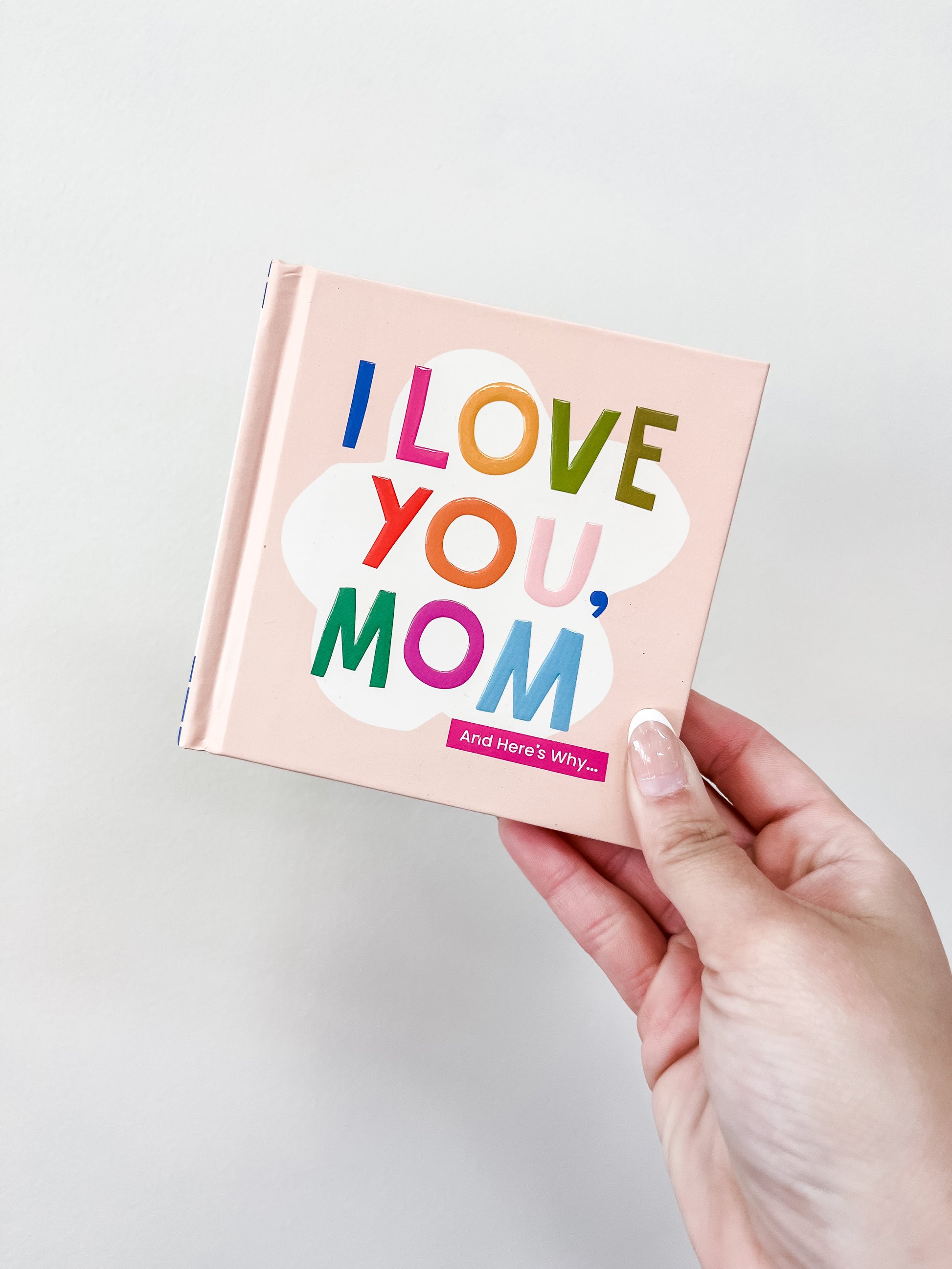 A hand holds the familuis I Love You, Mom Book featuring colorful lettering on a pink cover, set against a plain light background.