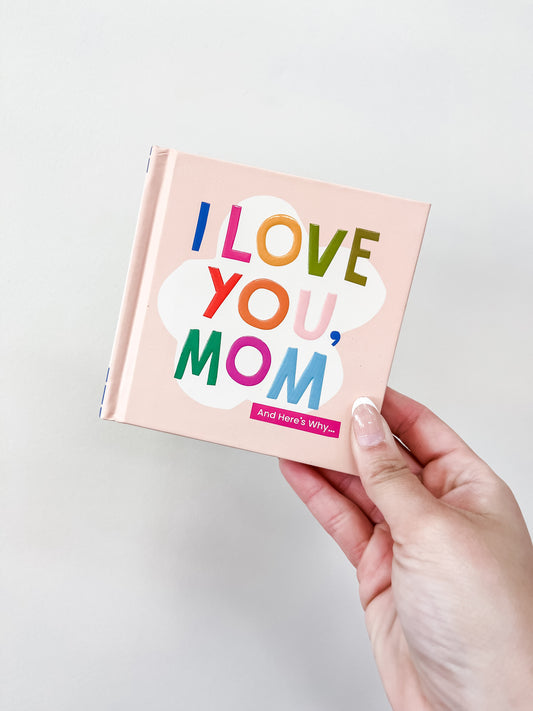A hand holds the familuis I Love You, Mom Book featuring colorful lettering on a pink cover, set against a plain light background.