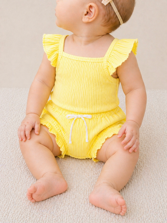 A baby in the Sunny Days Ribbed Set by Tiny Tot Bundles, featuring a yellow ruffled romper and headband, sits on a light textured surface with legs stretched out, looking slightly to the side.