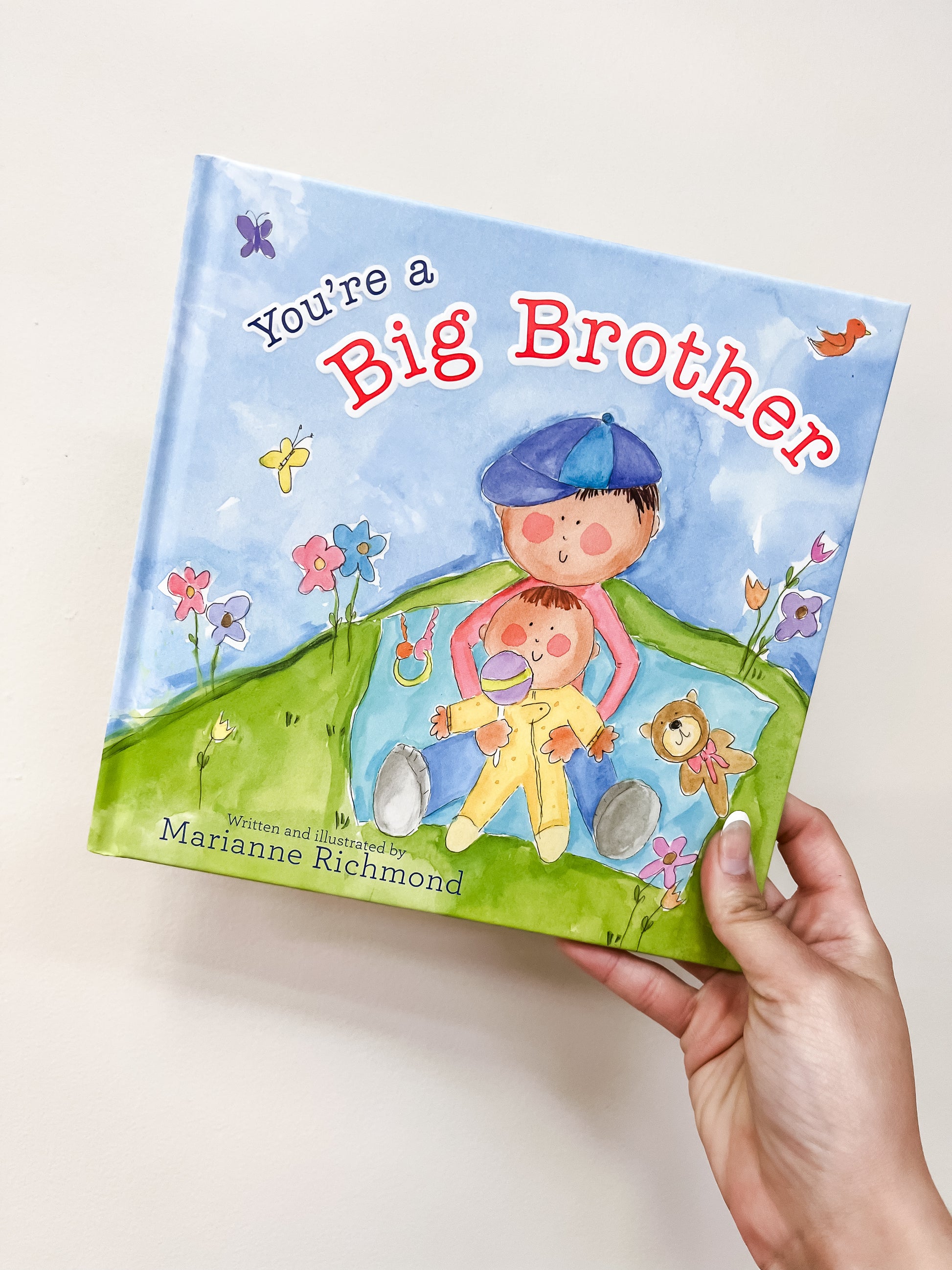 A hand holds the You're A Big Brother Book by Sourcebooks, showing an illustrated boy hugging a baby on a grassy hill with flowers, a teddy bear, and a blue sky in the background.