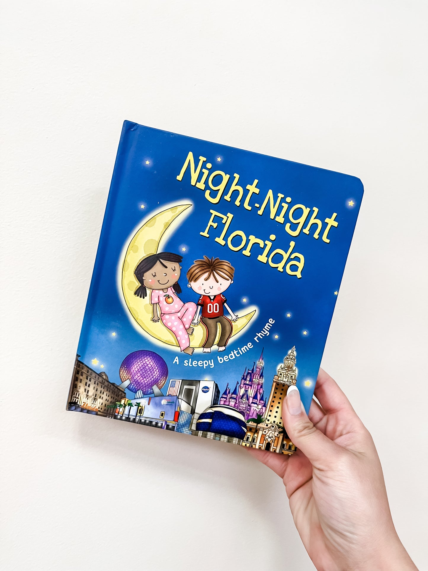 A hand holds the Night-Night Florida Book by Sourcebooks, showing illustrated children on a crescent moon above a cityscape beneath a starry sky.