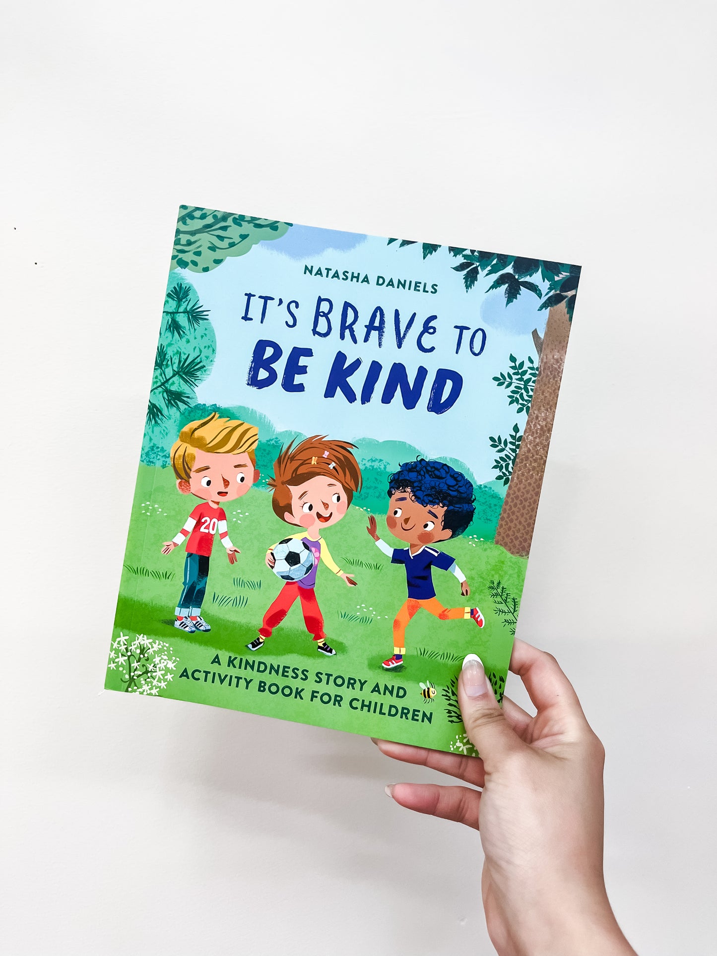 A hand holds the "It's Brave To Be Kind Book" by Sourcebooks, showing three children playing soccer on a grassy field.