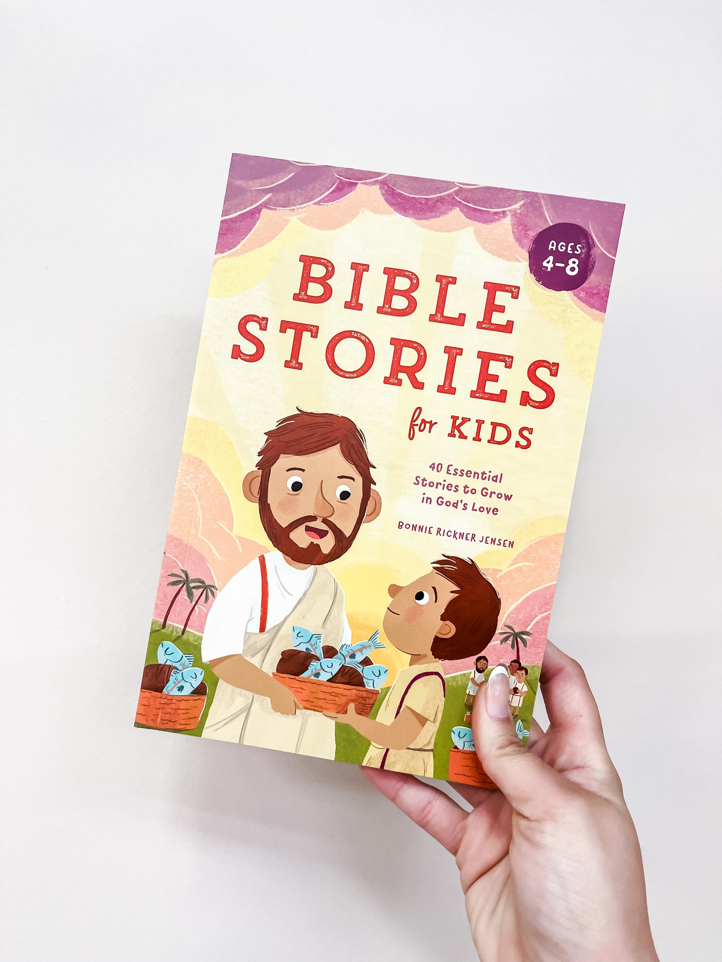 A hand holds the Bible Stories For Kids Book by Sourcebooks. The cover shows illustrated figures of a man and child with baskets of fish and bread.