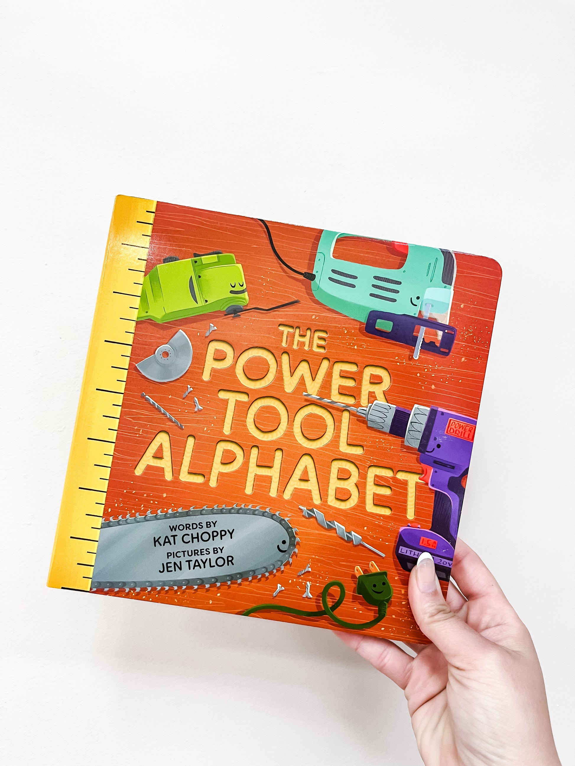 A hand holds the Power Tool Alphabet Book by Sourcebooks, a colorful children’s book with a bright orange cover featuring illustrated tools like a chainsaw, drill, circular saw blade, and jigsaw.