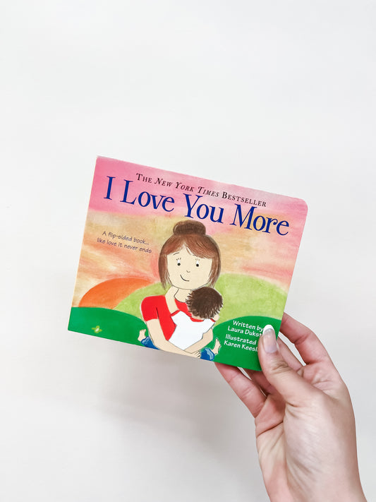 A hand holds the I Love You More Book by Sourcebooks, featuring a cover with a woman hugging a child against a pastel background.