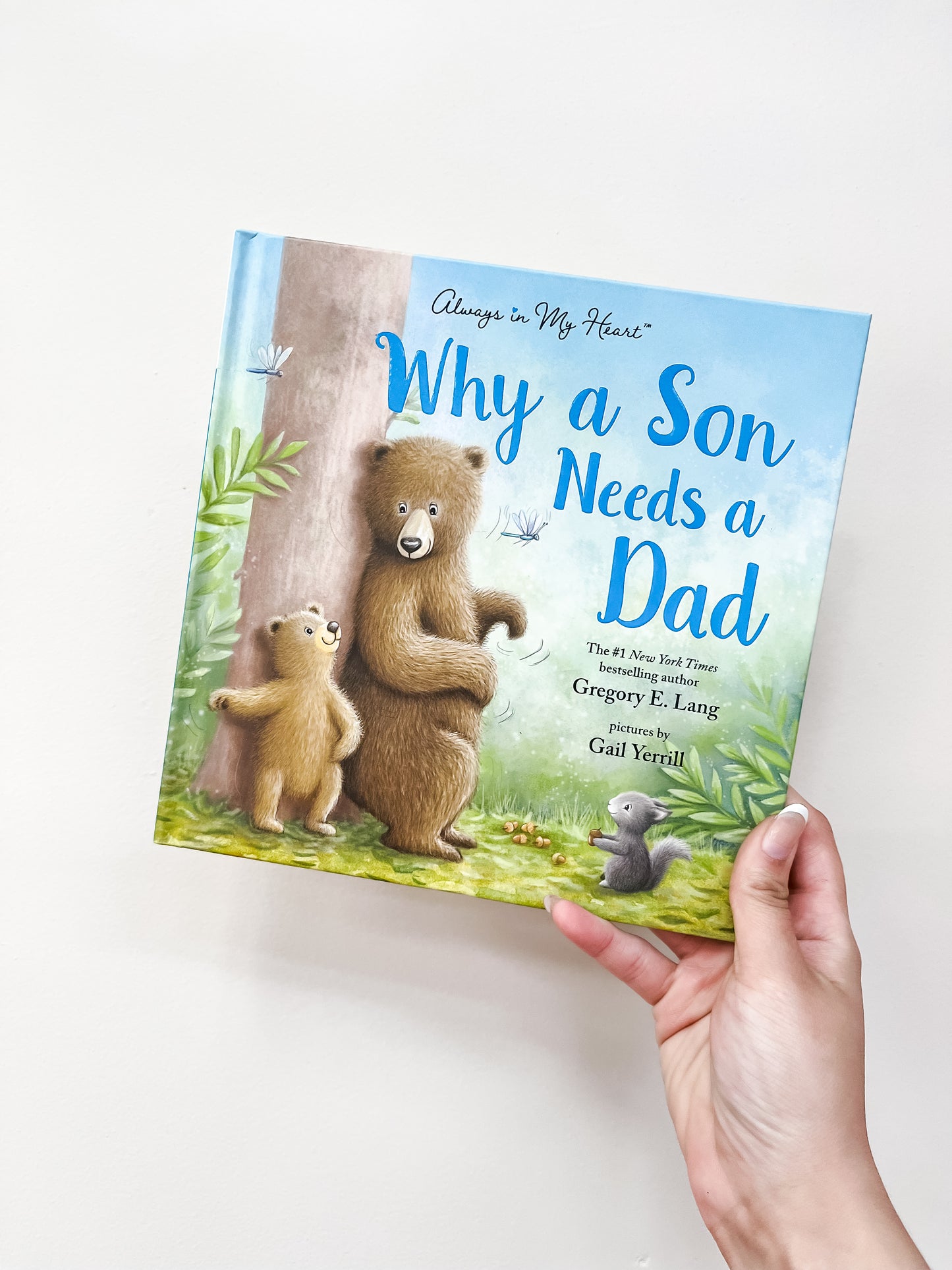 A hand holds the "Why A Son Needs A Dad Book" by sour, which features two bears—an adult and a cub—waving next to a small gray bird on the cover.