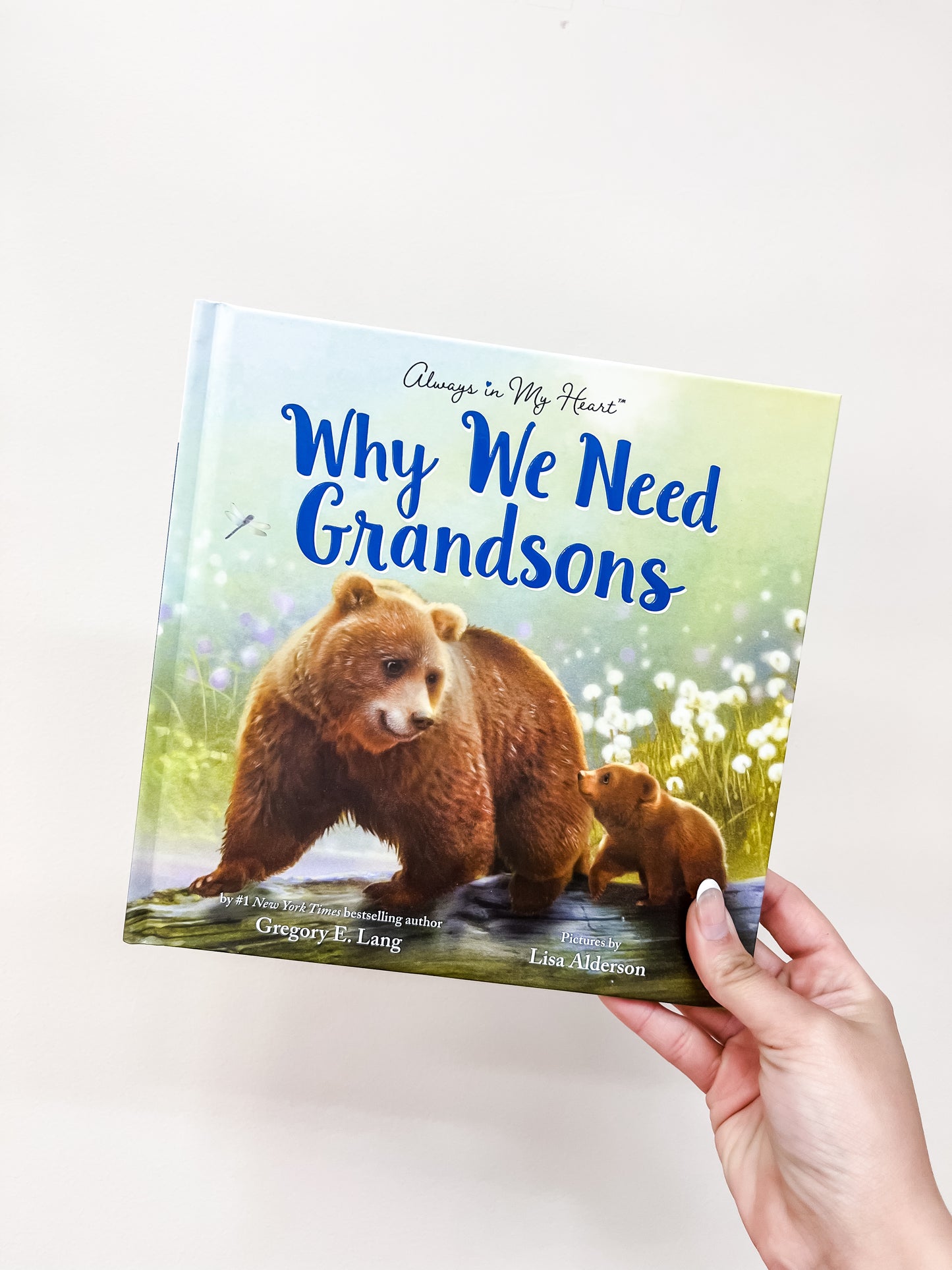A hand holds the Why We Need Grandsons Book by sour, featuring a cover illustration of a bear and her cub sitting together in a grassy, flower-filled field.