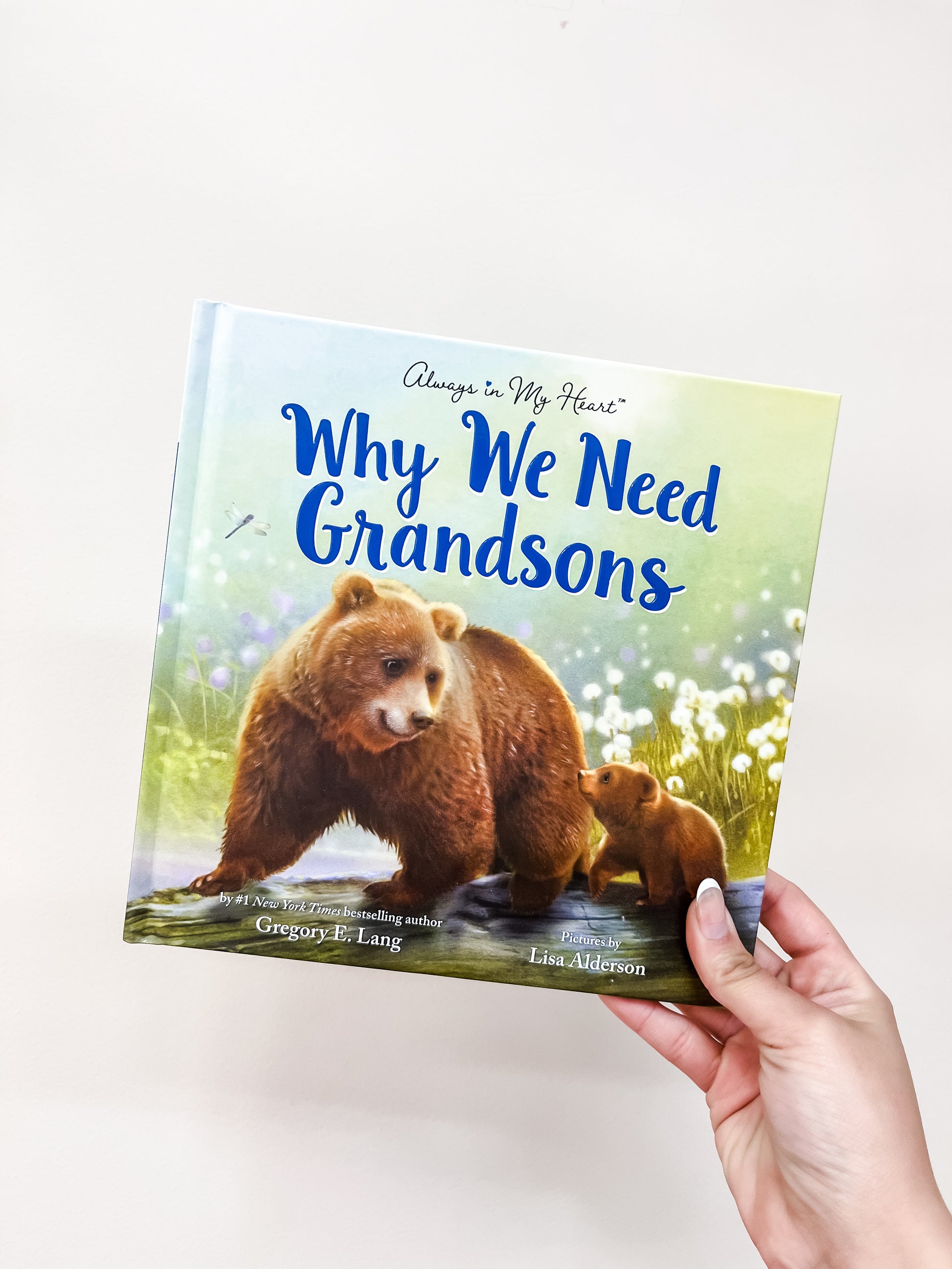 A hand holds the Why We Need Grandsons Book by sour, featuring a cover illustration of a bear and her cub sitting together in a grassy, flower-filled field.