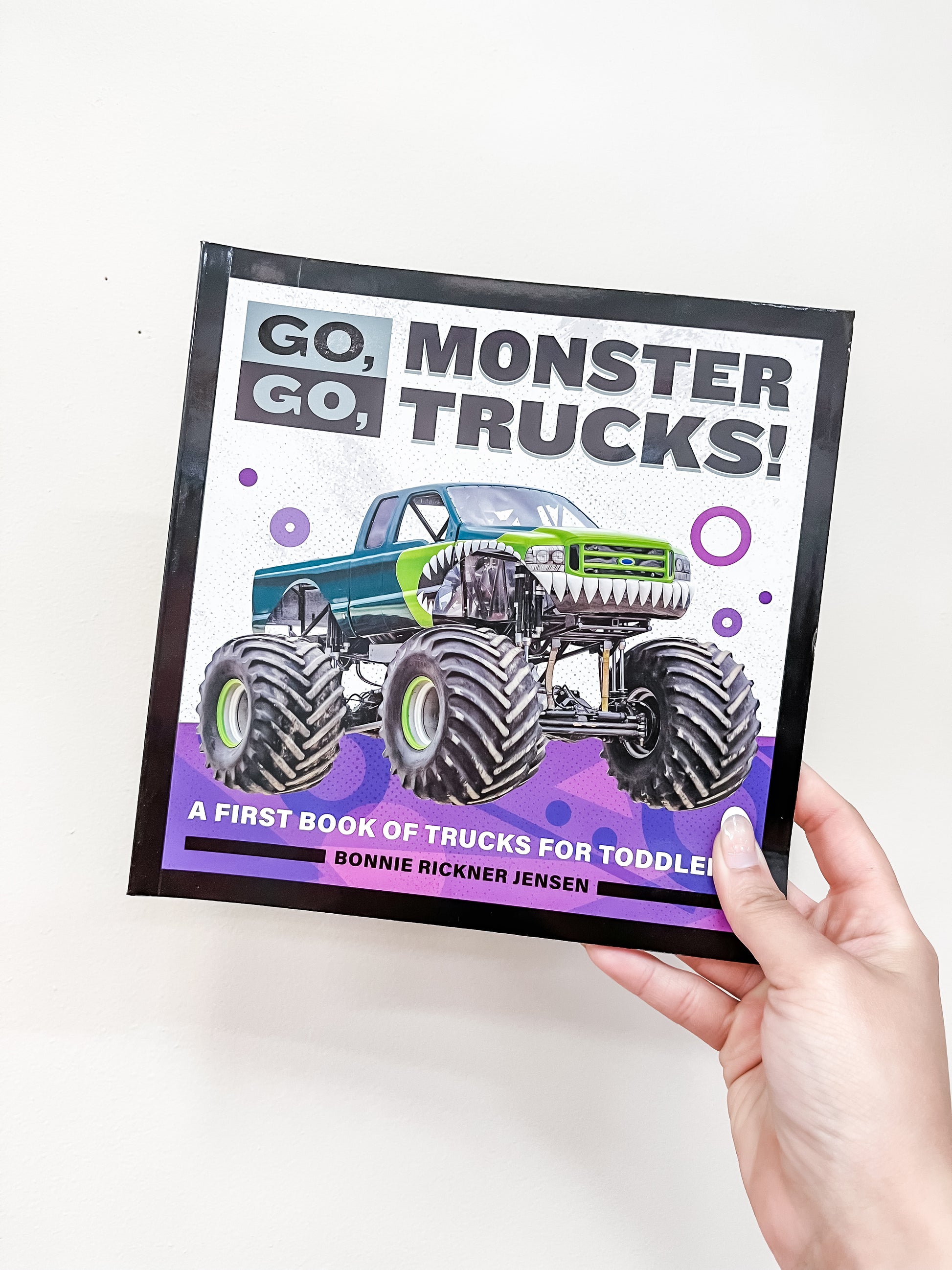 A hand holds the Go, Go, Monster Trucks! Book by Sourcebooks, a children's book featuring an illustrated green monster truck on the cover.