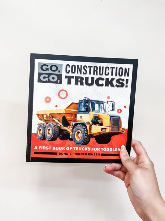 A hand holds the Go, Go, Construction Trucks Book by Sourcebooks, a children's book for toddlers featuring a yellow dump truck on the cover.