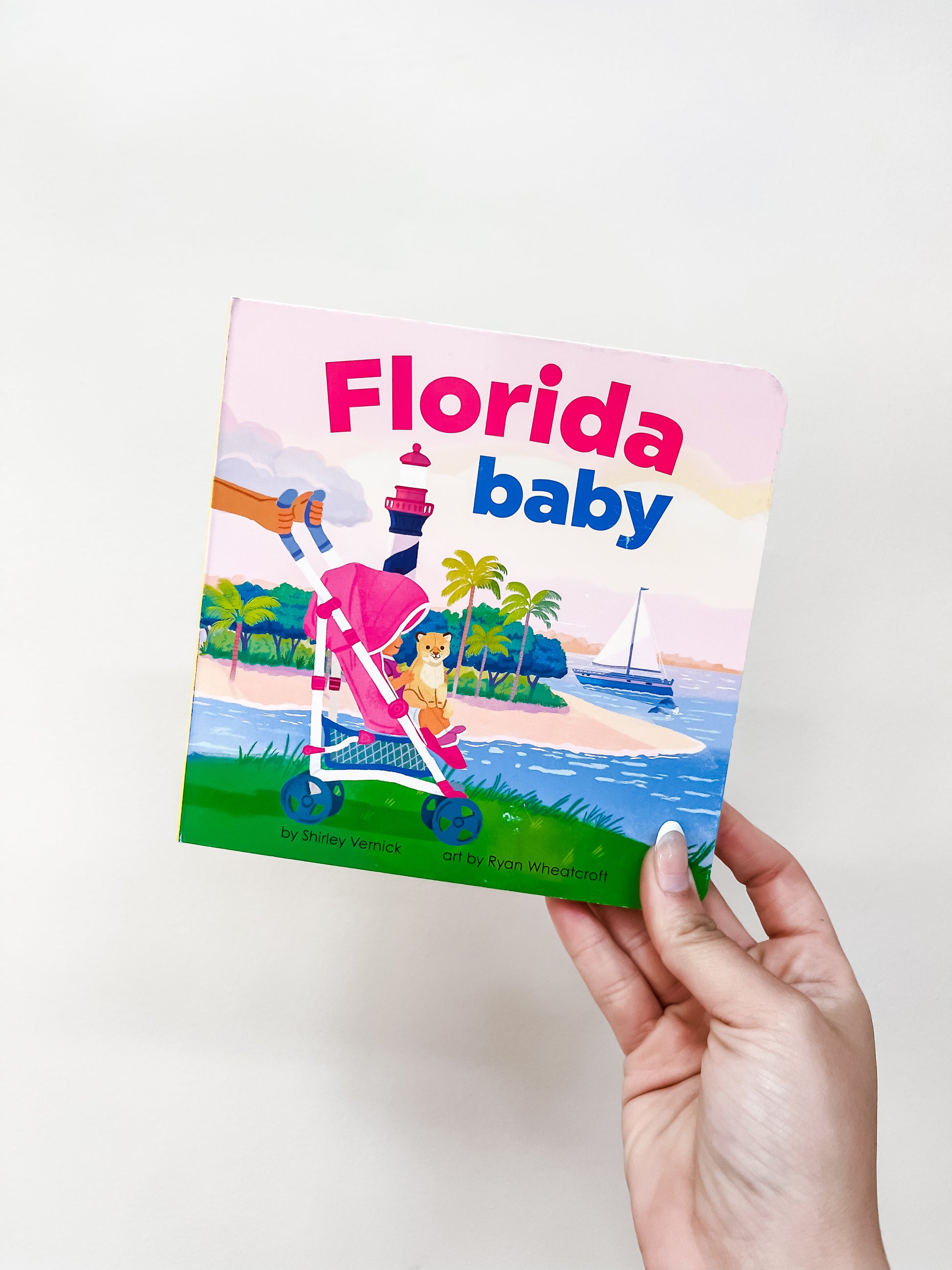 A hand holds the Florida Baby Book by Sourcebooks, featuring a baby in a stroller at the beach with palm trees, a sailboat, and a lighthouse in the background.