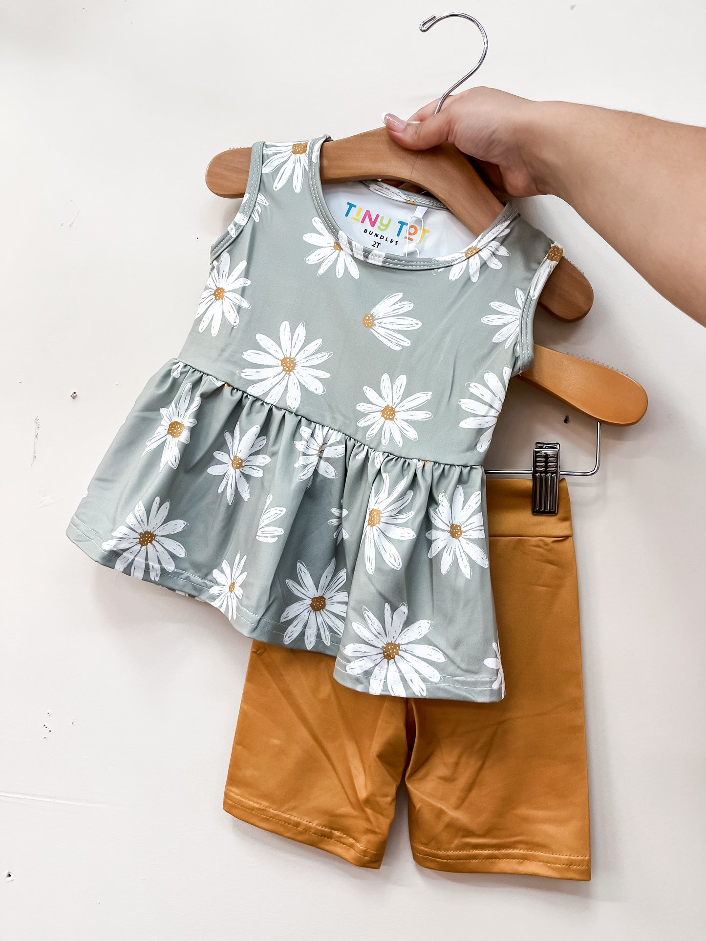 A hand displays the Daisy Daydream Peplum Set by Tiny Tot Bundles: a sleeveless green top with white daisy prints and ruffled hem, paired with mustard yellow shorts.