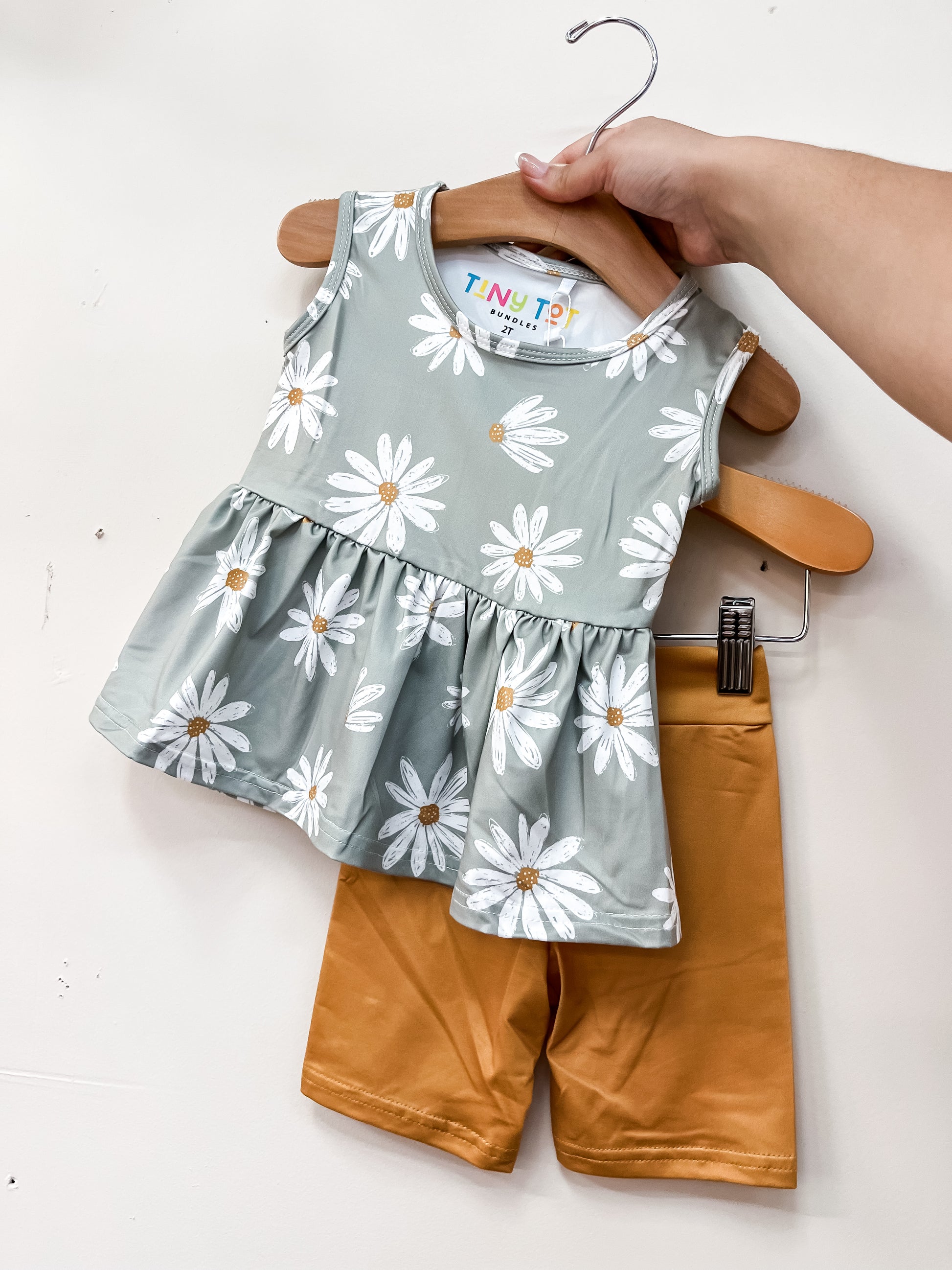 A hand displays the Daisy Daydream Peplum Set by Tiny Tot Bundles: a sleeveless green top with white daisy prints and ruffled hem, paired with mustard yellow shorts.