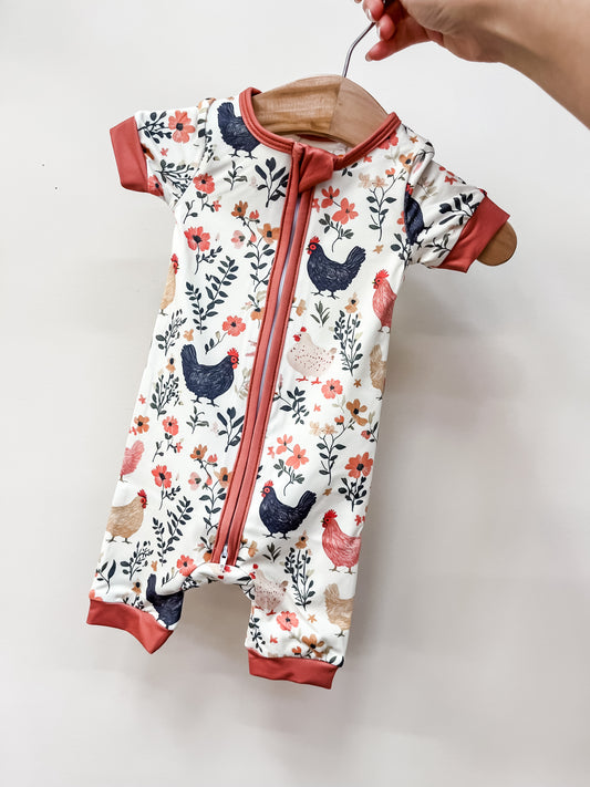 A hand holds a Tiny Tot Bundles Farmyard Florals Shorty, a short-sleeved baby romper with colorful chicken and floral patterns, and rust-colored trim on the sleeves, cuffs, and zipper.