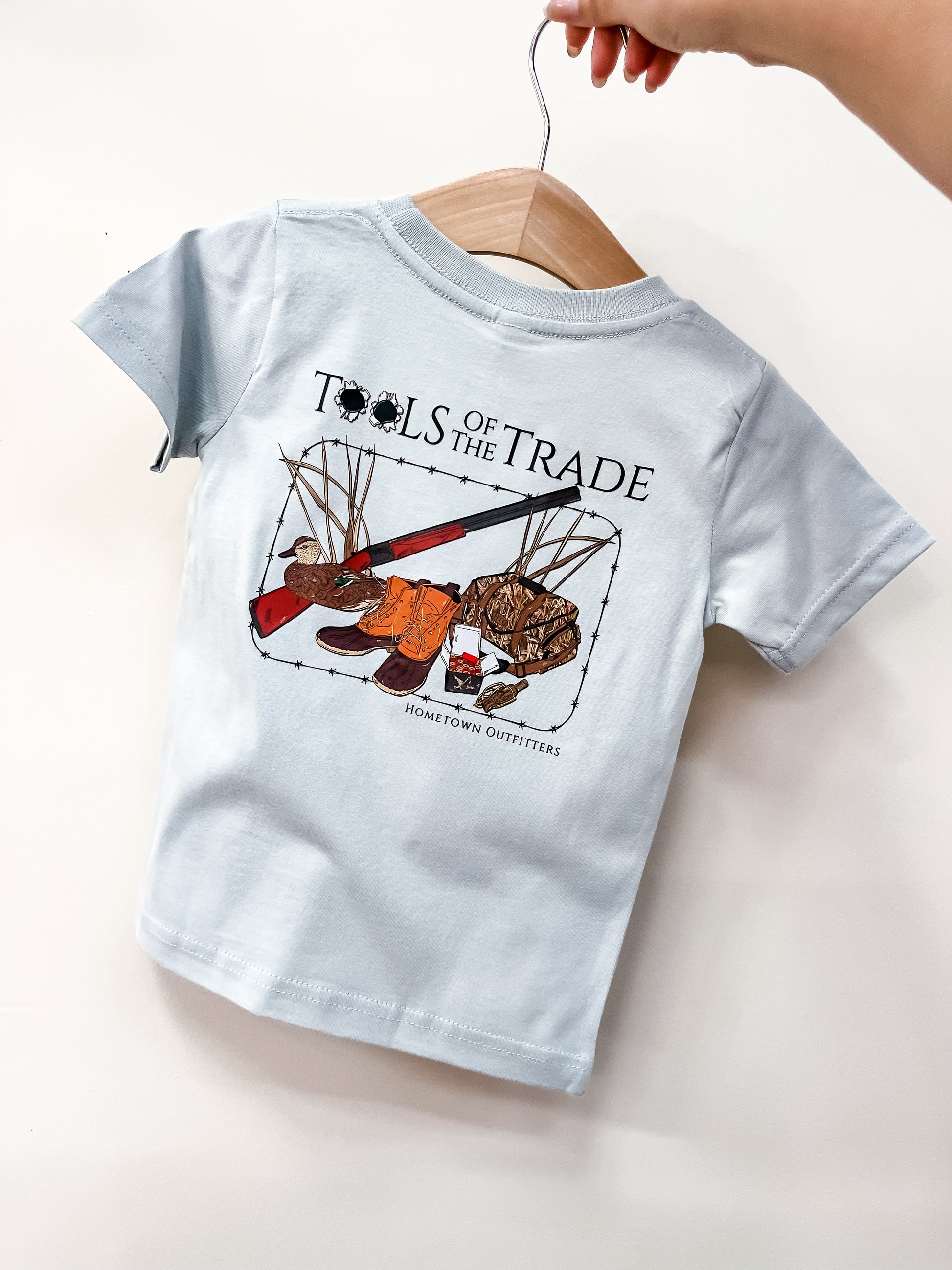 A hand holds a light blue Tools Of The Trade T-Shirt by hometown Outfitters on a wooden hanger. The back features hunting gear graphics—boots, gloves, duck decoys, shotgun—with "TOOLS OF THE TRADE" and "hometown Outfitters" text.