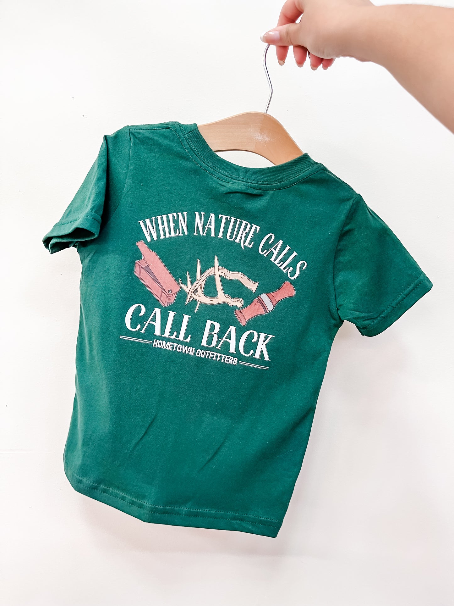 A hand holds a green Nature Calls T-Shirt by hometown Outfitters on a wooden hanger. The back features "WHEN NATURE CALLS, CALL BACK." and graphics of logs, an antler, and a flashlight.