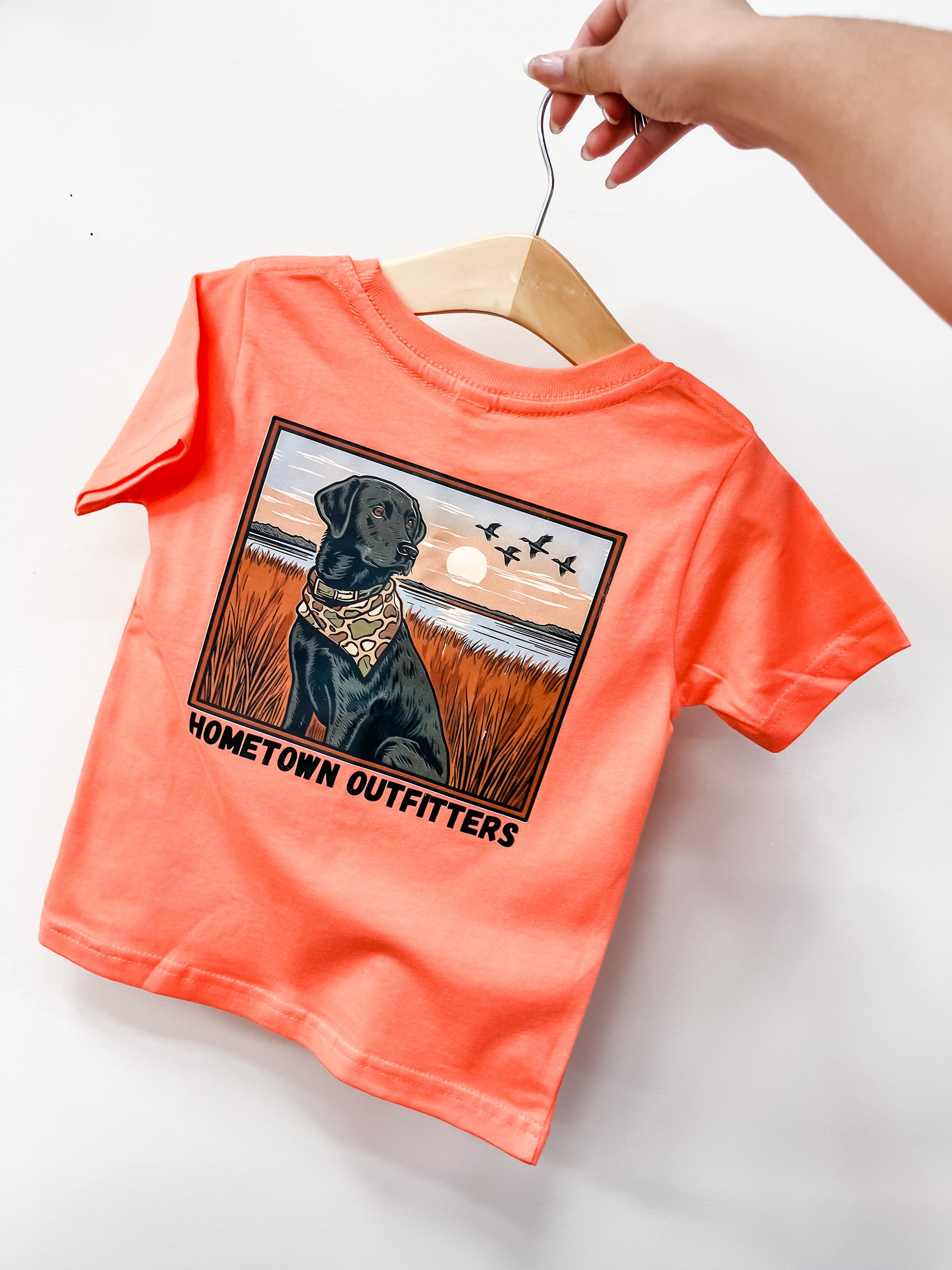 A hand holds the coral “Sunset Retrieve T-Shirt” by hometown Outfitters, featuring a black dog in a bandana sitting in a grassy field at sunset with flying birds and the text “HOMETOWN OUTFITTERS.”.